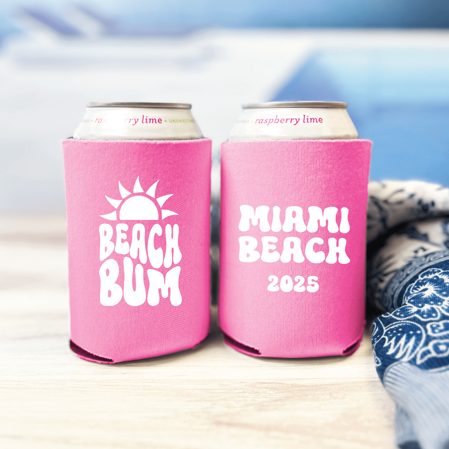 Two pink neoprene can coolers with white text reading "Beach Bum" and "Miami Beach 2025" displayed with cans on a beach setting.