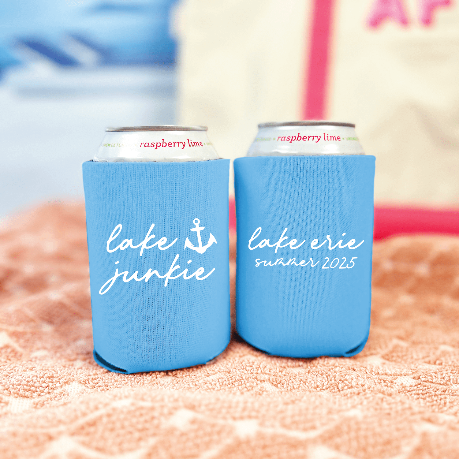 Pair of bright blue neoprene can coolers with "lake junkie" and "cafe erie summer 2025" text and anchor design, holding raspberry lime cans on sandy beach.