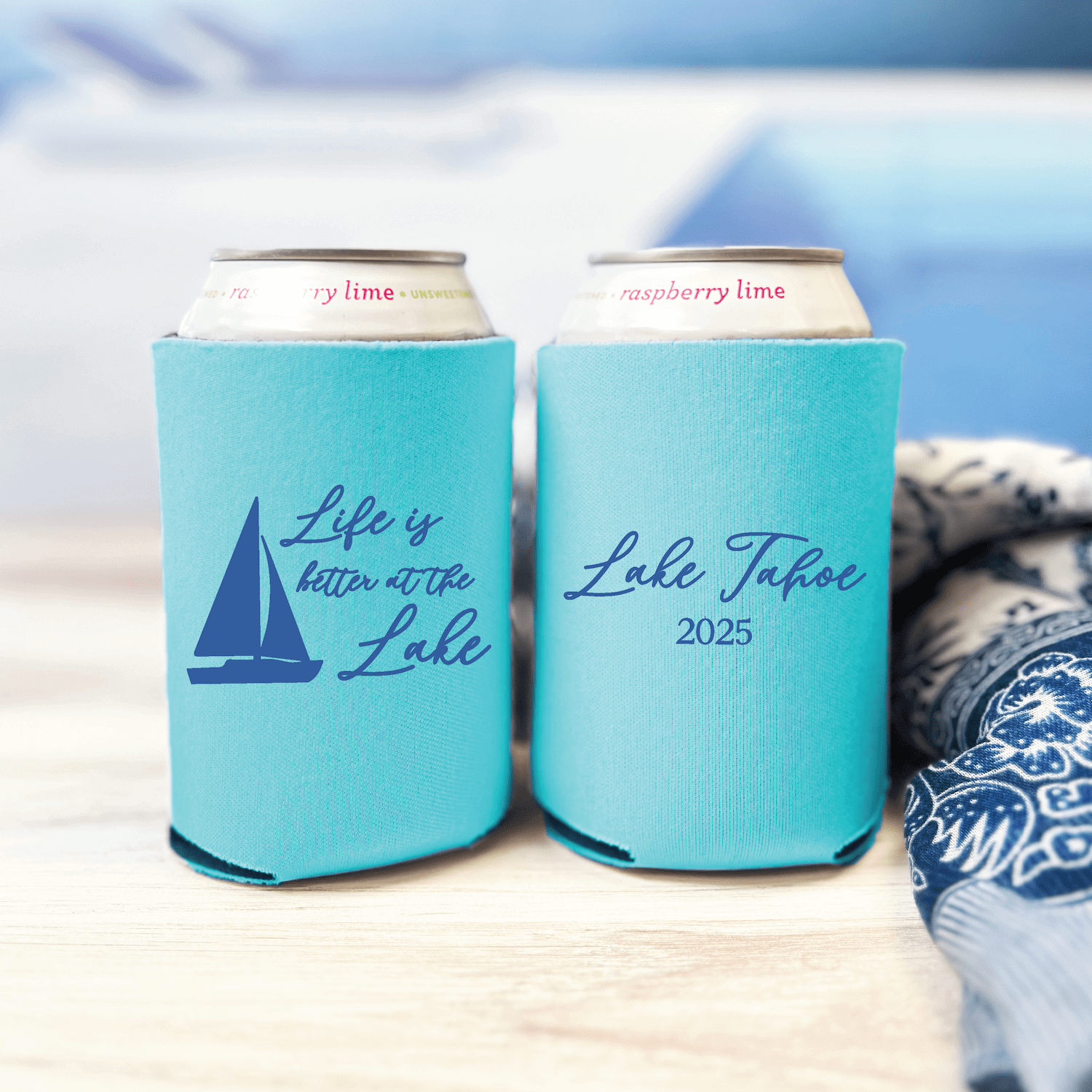 Turquoise can coolers with sailboat design and personalized "Lake Tahoe 2025" text, displayed side by side on wooden surface