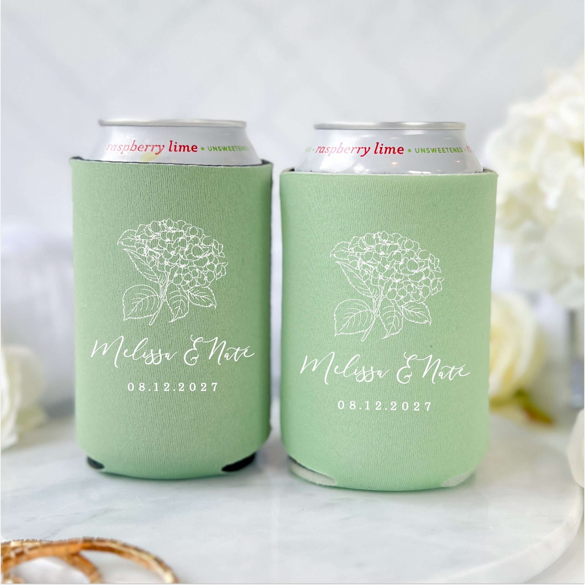Two personalized sage green fabric can coolers feature a delicate white outline drawing of a hydrangea flower, ideal for keeping beverages cold at a wedding or special event.