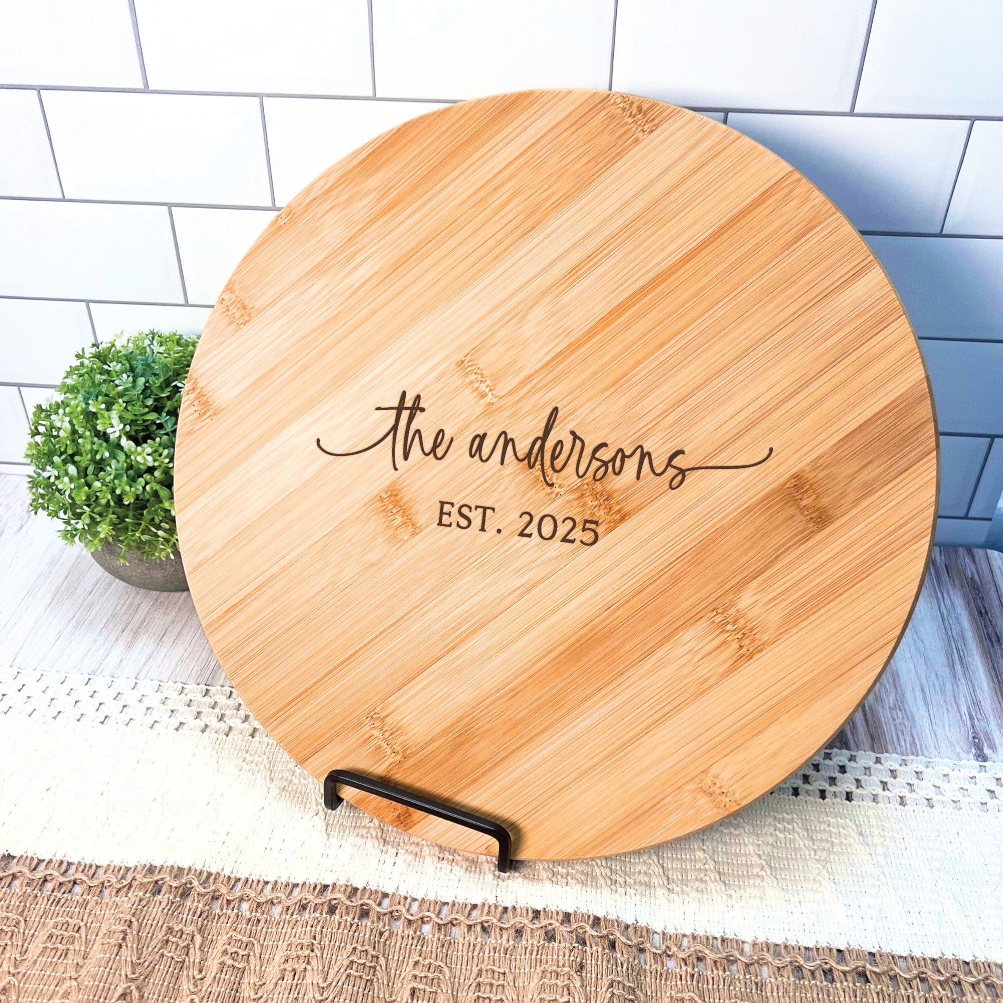 A round personalized bamboo serving board with laser-engraved text, displayed on a black metal stand, perfect as a wedding gift or home decor item.