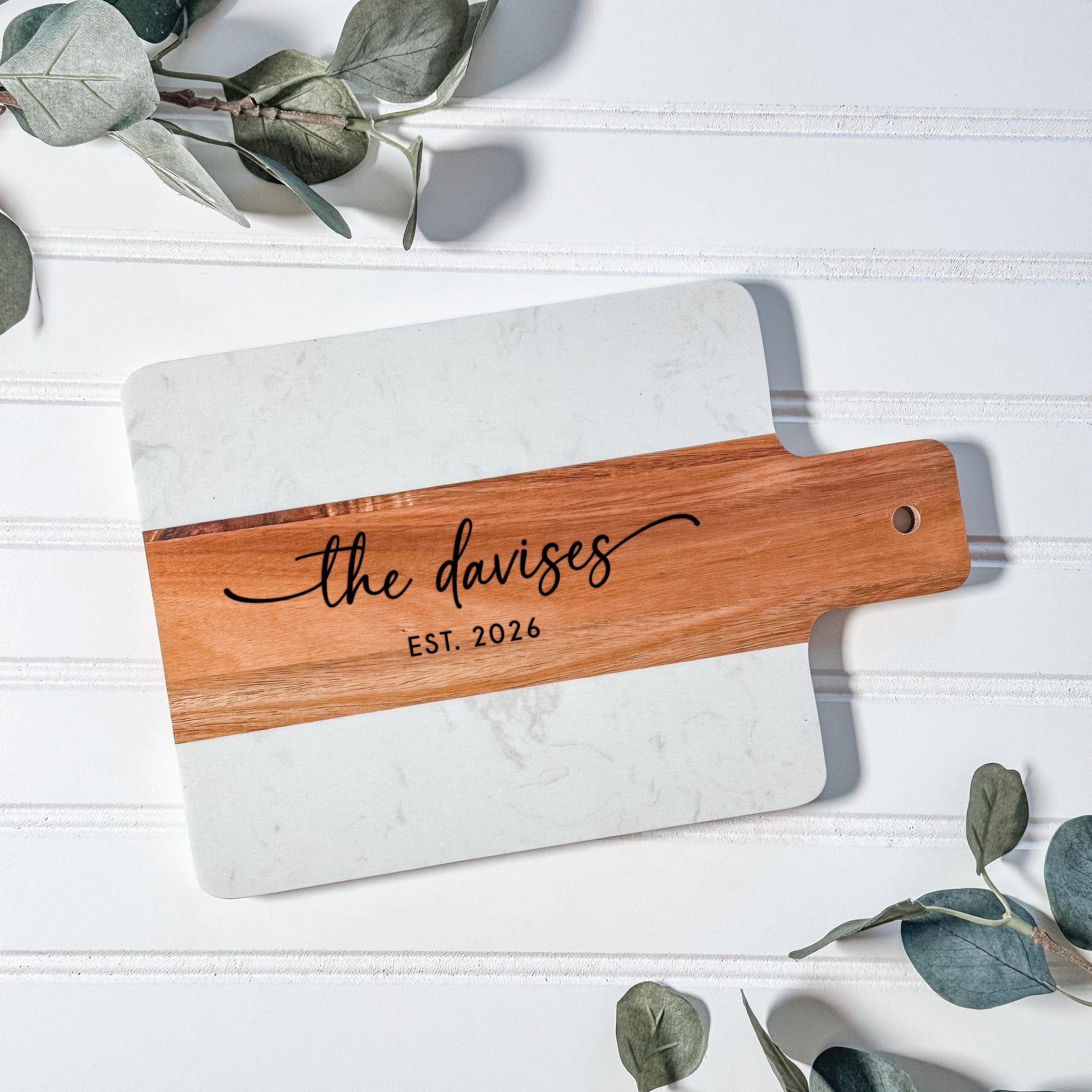 A rectangular personalized wedding or housewarming serving board crafted from white marble and a contrasting light brown wooden strip with a handle.