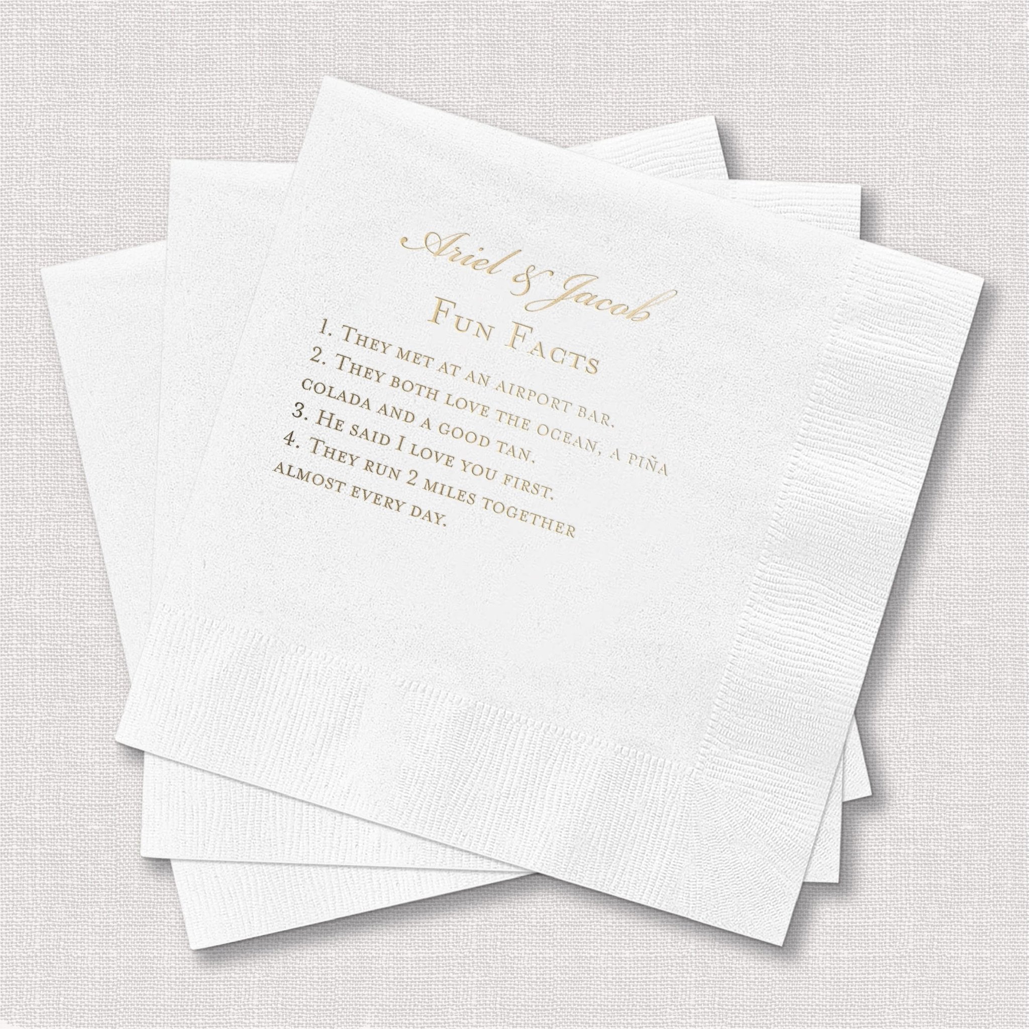 White cocktail napkins with gold foil stamped text reading "About Our Love" and fun facts about the couple
