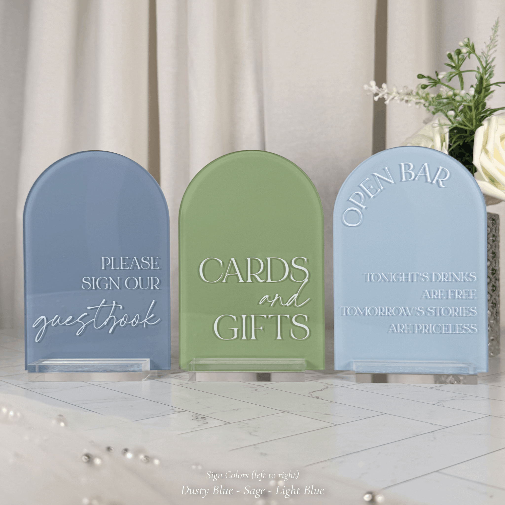 Three arched acrylic wedding signs in dusty blue, sage green, and light blue on clear stands with fresh florals in the background, reading Open Bar Tonight's Drinks Are Free Tomorrow's Stories Are Priceless, Please Sign Our Guestbook, and Cards and Gifts.