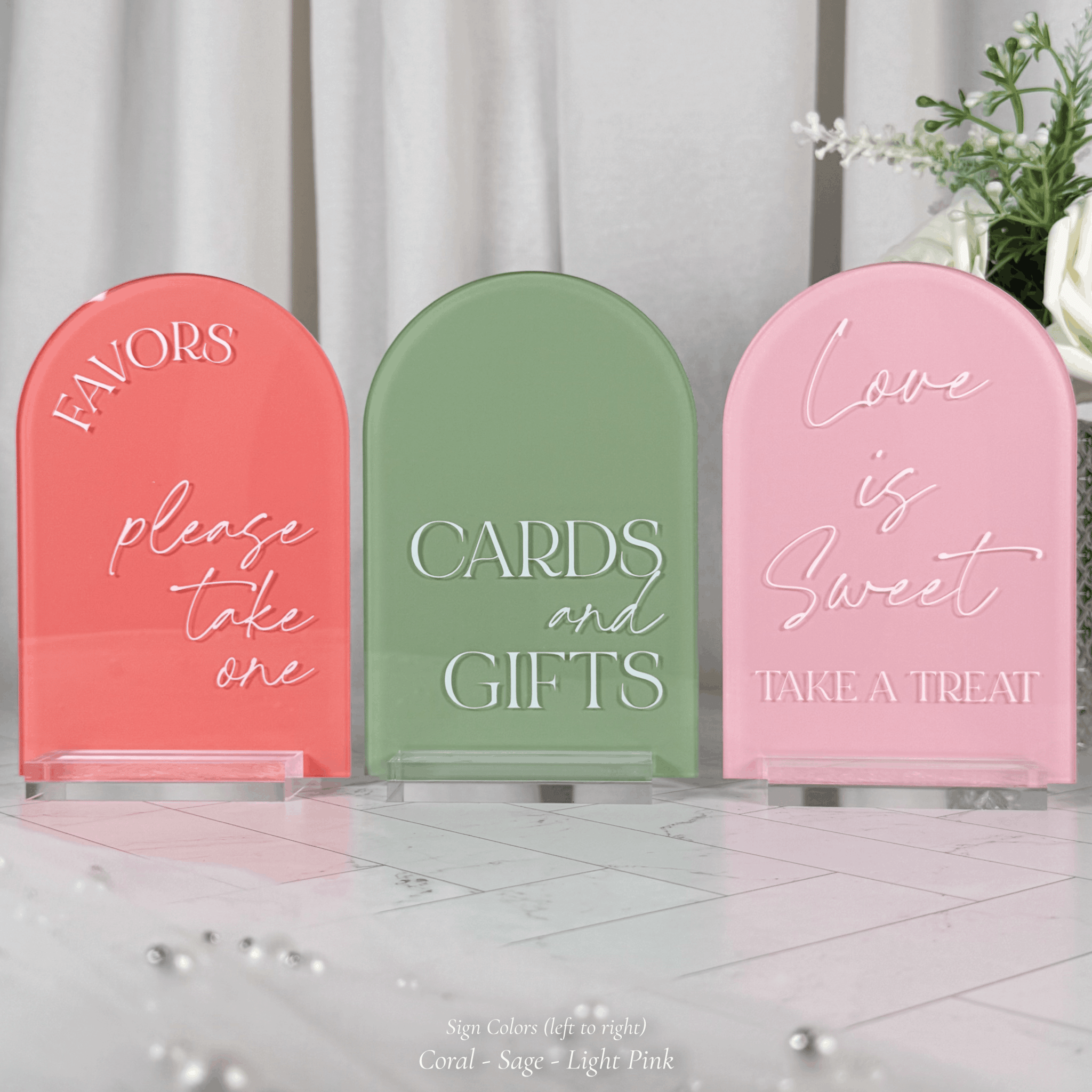 Three arched acrylic wedding signs in coral, sage green, and light pink on clear stands with fresh florals in the background, reading Favors Please Take One, Cards and Gifts, and Love is Sweet Take a Treat.