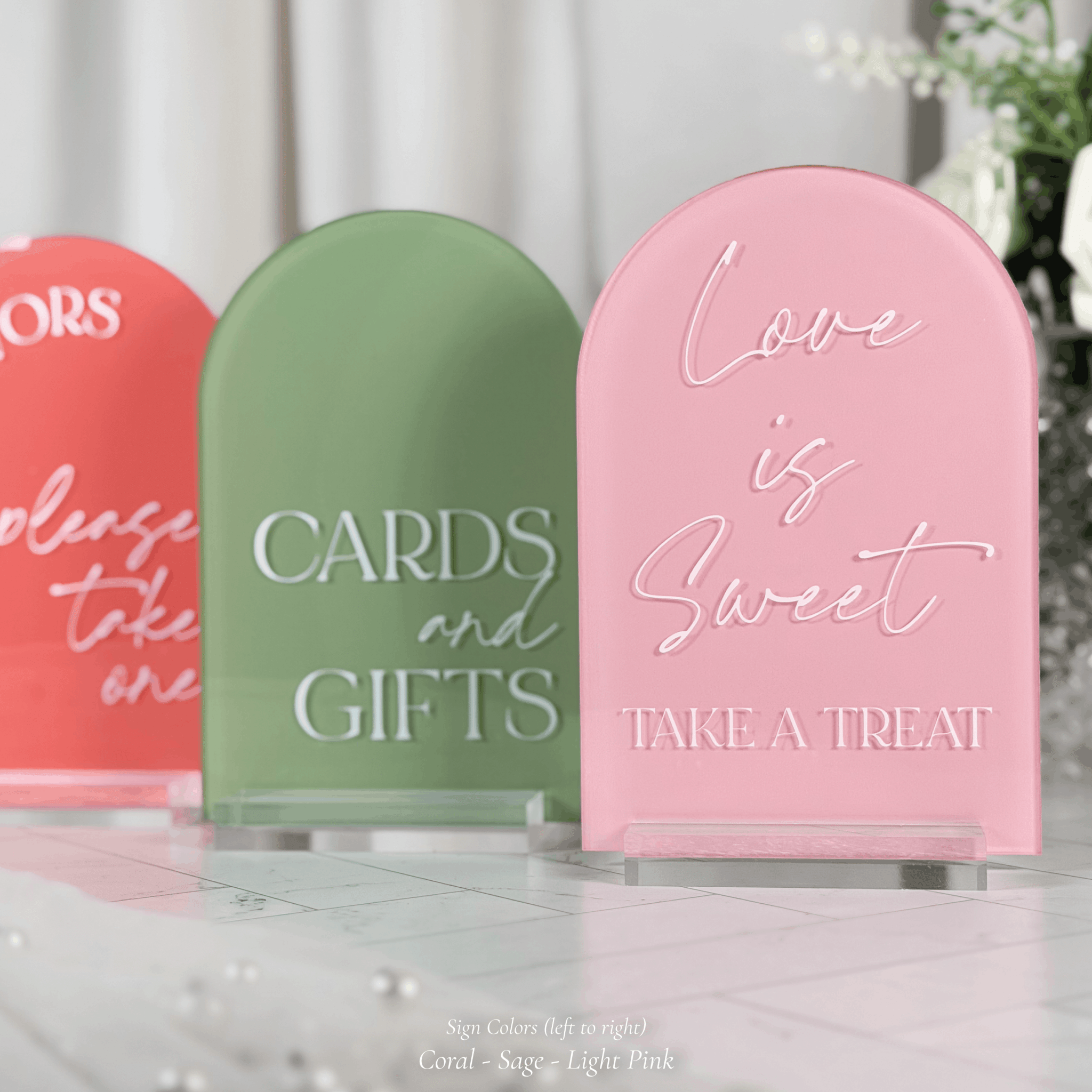 Close-up of three arched acrylic wedding signs in coral, sage green, and light pink on clear stands, reading Favors Please Take One, Cards and Gifts, and Love is Sweet Take a Treat.