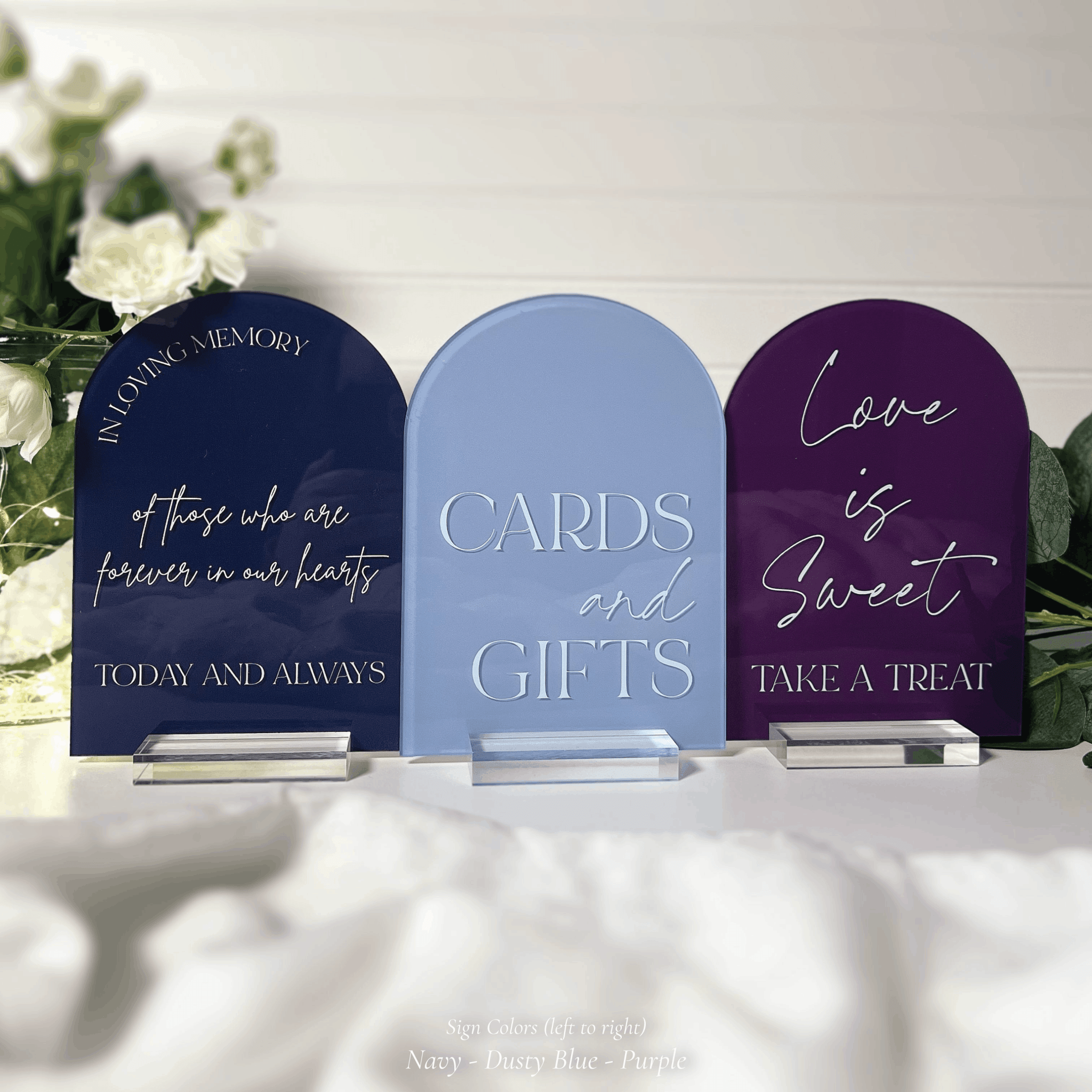 Three arched acrylic wedding signs in navy, dusty blue, and purple on clear stands with white florals in the background, reading In Loving Memory of Those Who Are Forever in Our Hearts Today and Always, Cards and Gifts, and Love is Sweet Take a Treat.