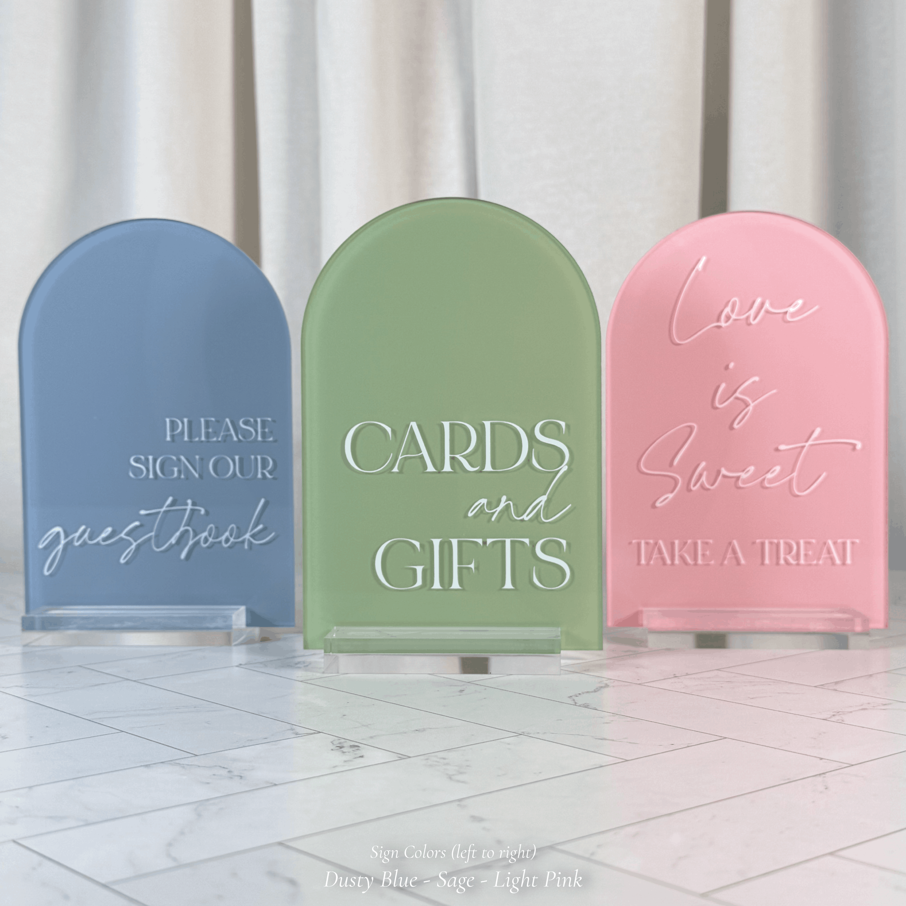 Three arched acrylic wedding signs in dusty blue, sage green, and light pink on clear stands atop a marble floor with a flowing curtain backdrop, reading Please Sign Our Guestbook, Cards and Gifts, and Love is Sweet Take a Treat.