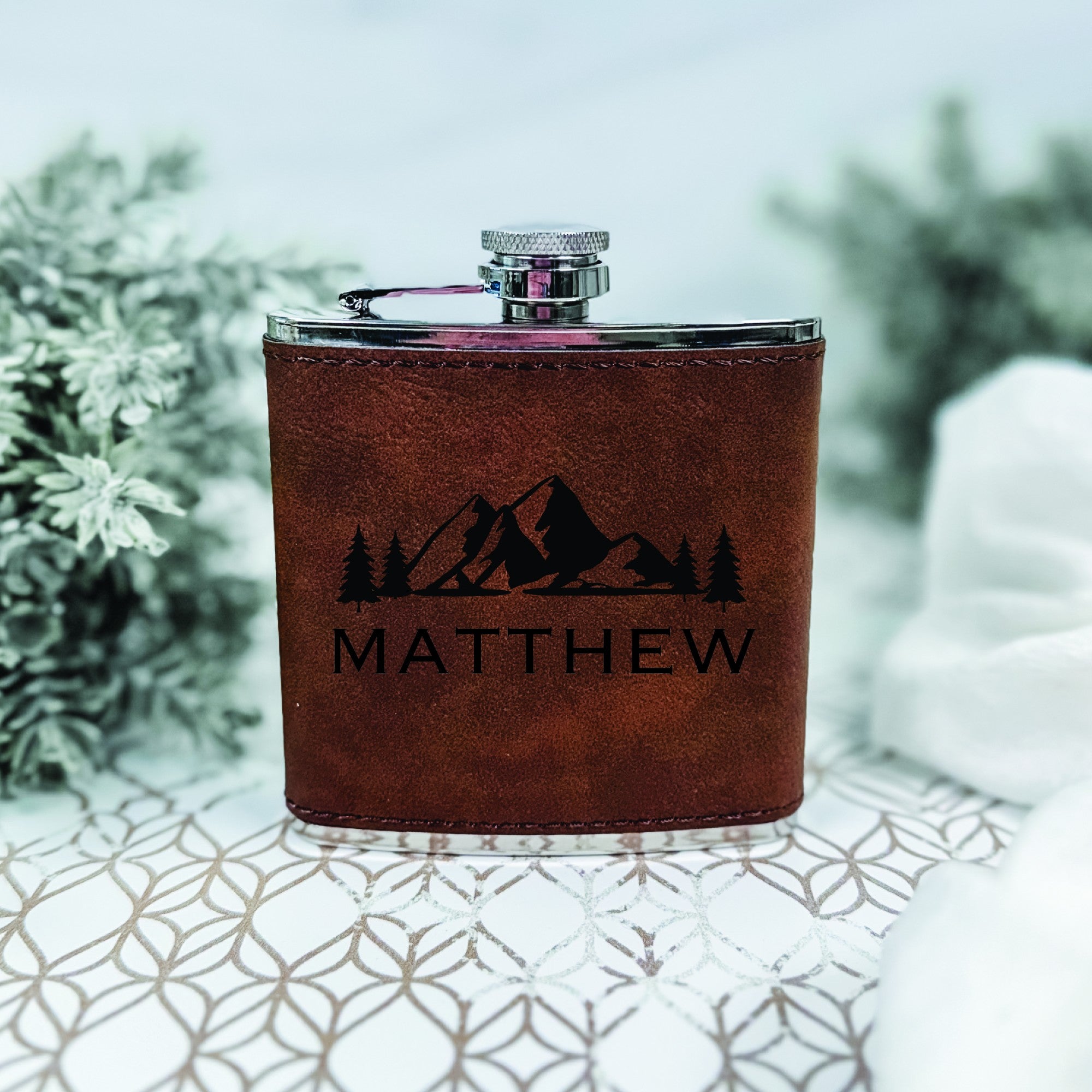 A brown leather-wrapped stainless steel hip flask, featuring an engraved mountain and pine tree design, suitable as a personalized gift for weddings or special events.