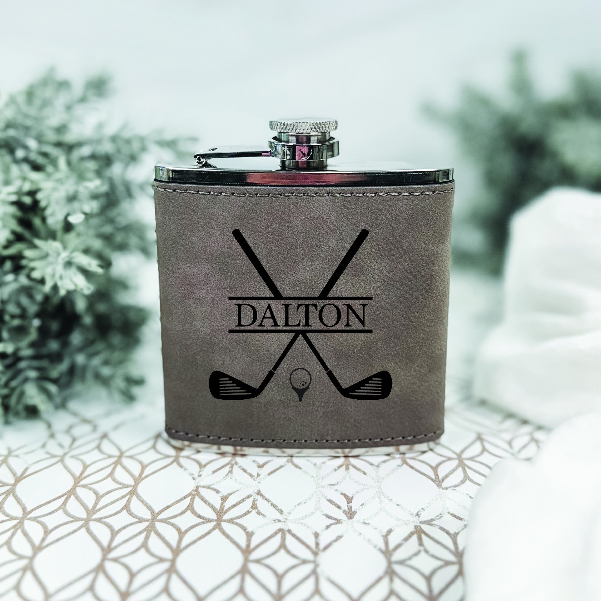 A gray leatherette hip flask with a silver screw cap, featuring an engraved design of crossed golf clubs and a golf ball on a tee, ideal as a personalized gift for a golf enthusiast.