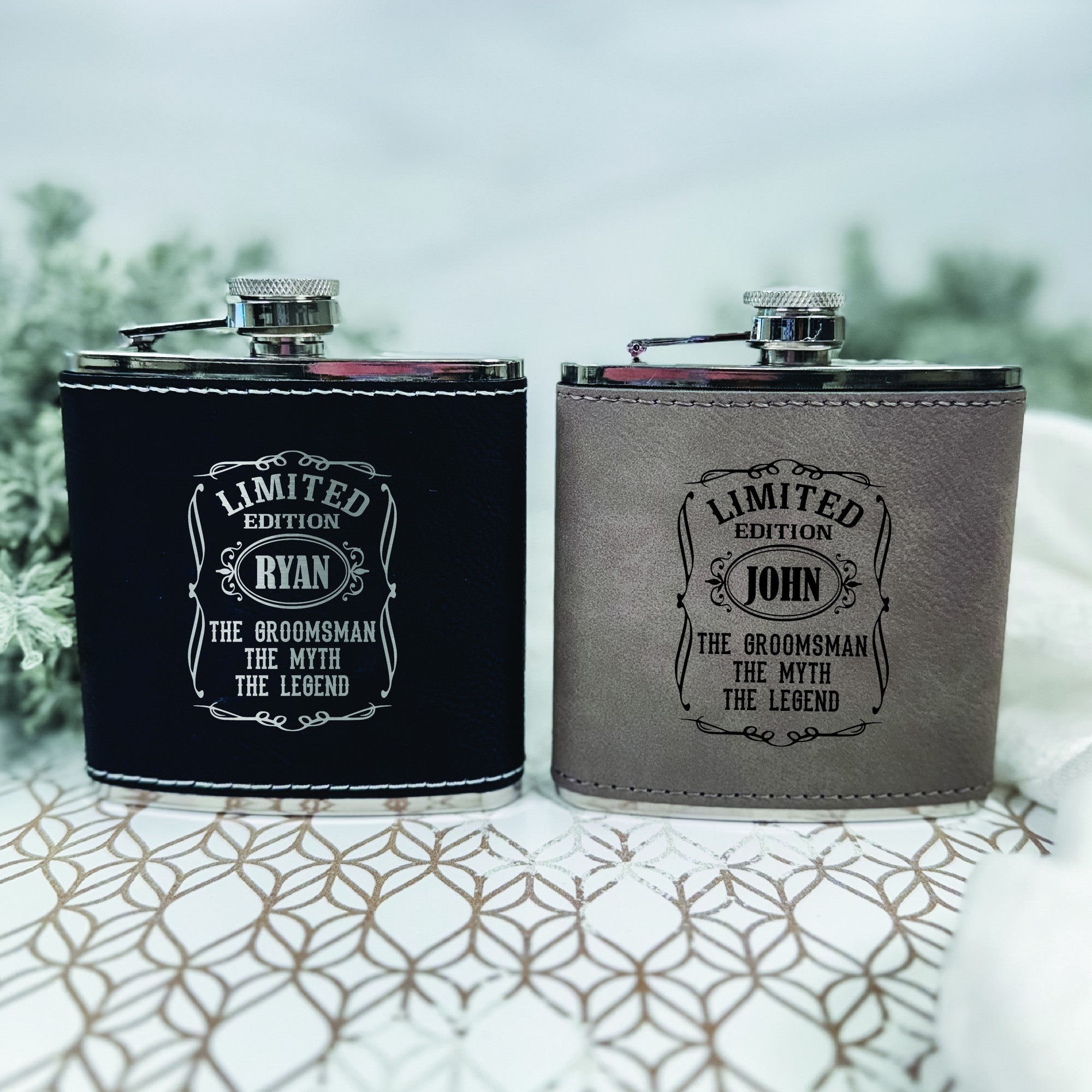 Two personalized hip flasks, one black and one grey faux leather-wrapped stainless steel, featuring custom engravings, presented as groomsmen gifts for a wedding.