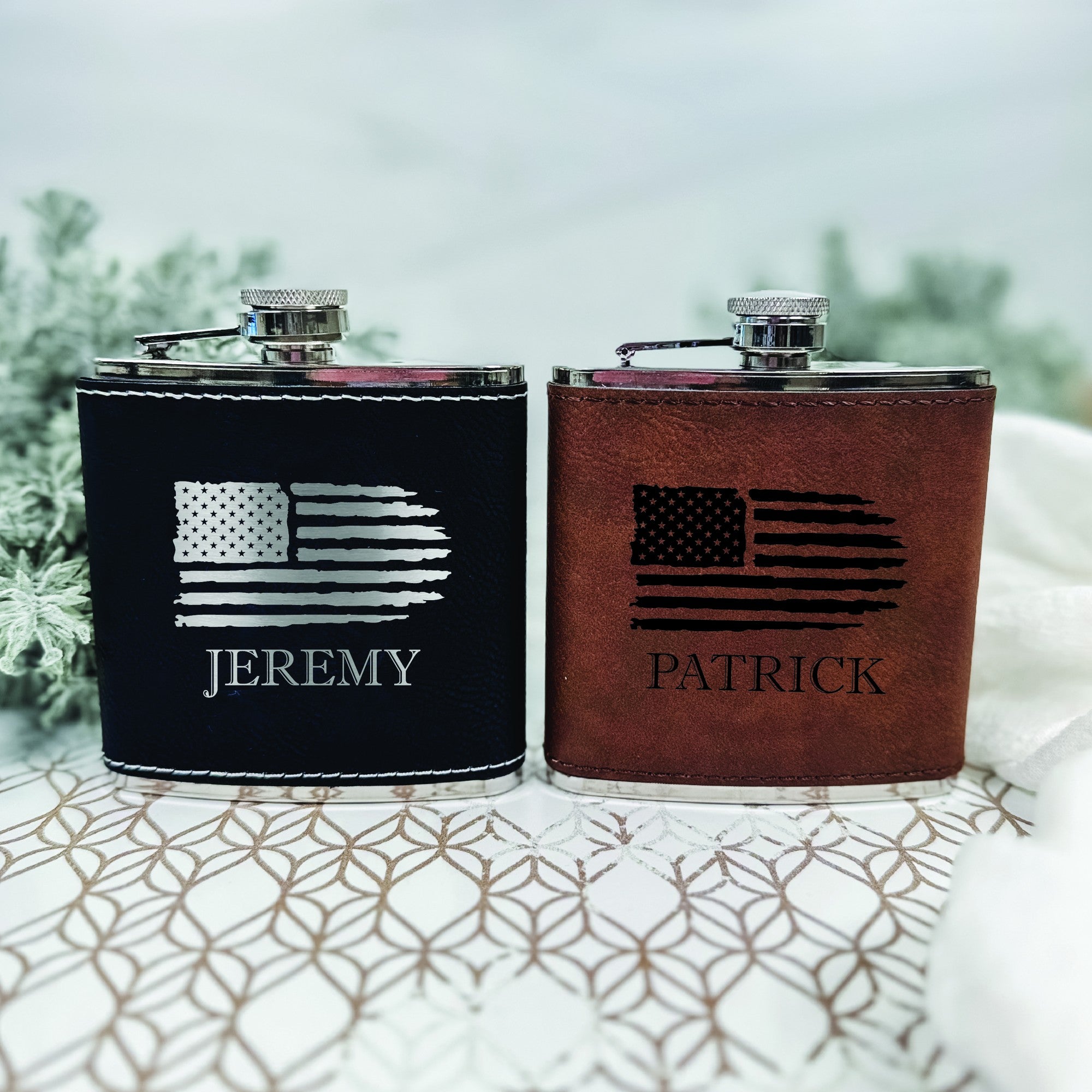 A pair of stainless steel flasks, one black and one brown, each wrapped in faux leather and engraved with a distressed American flag design, topped with a shiny silver screw cap, for a personalized gift.