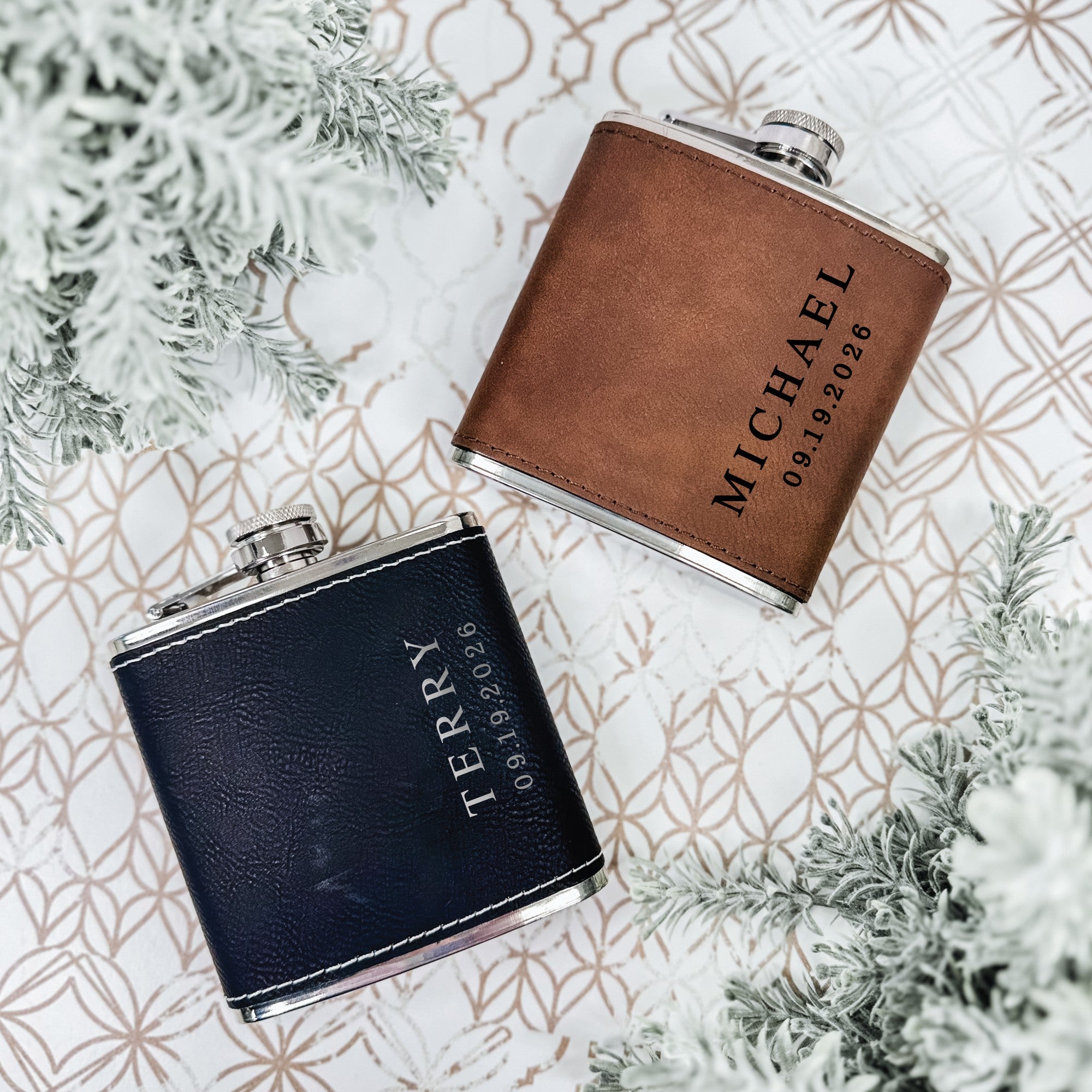 Two rectangular stainless steel hip flasks are visible, one wrapped in brown leatherette and the other in black leatherette, suitable as personalized gifts for a wedding or special event.