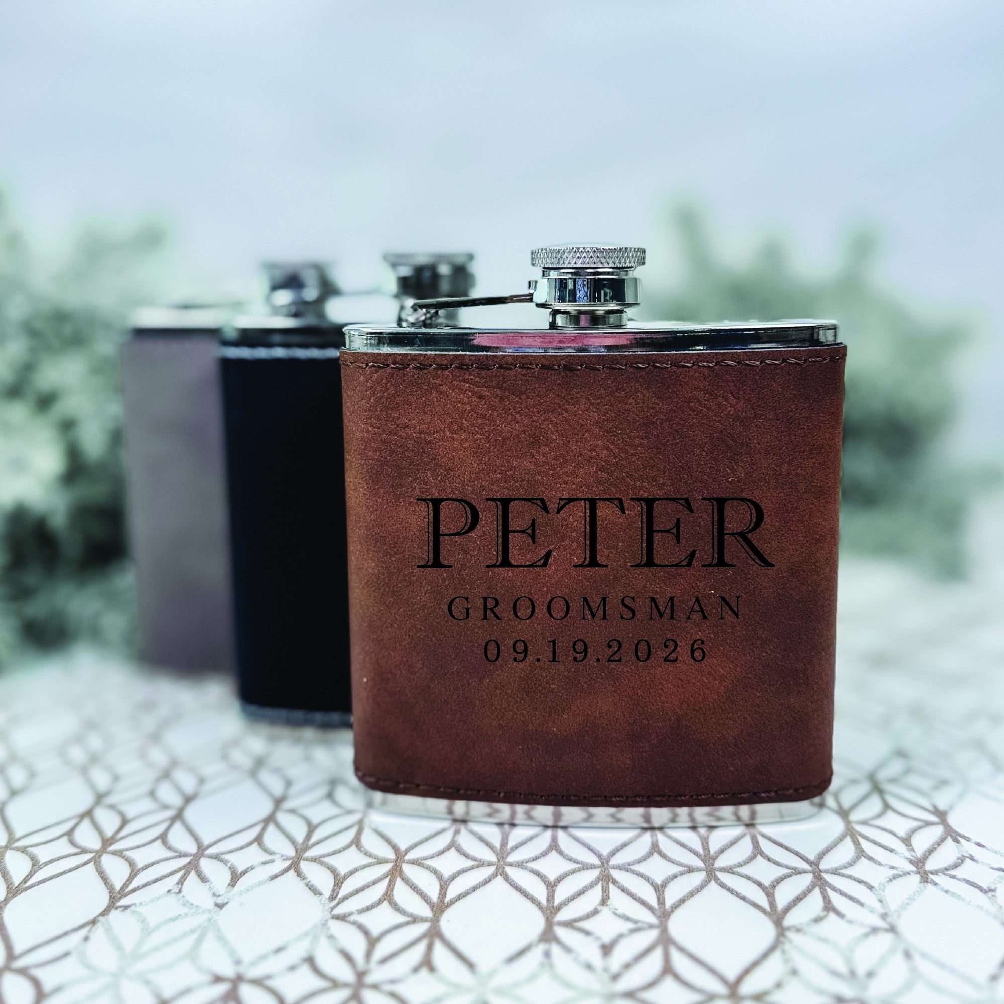 A brown leather-wrapped stainless steel flask with a screw-top lid stands in the foreground, accompanied by two blurred flasks in grey and black, ideal as personalized wedding gifts.