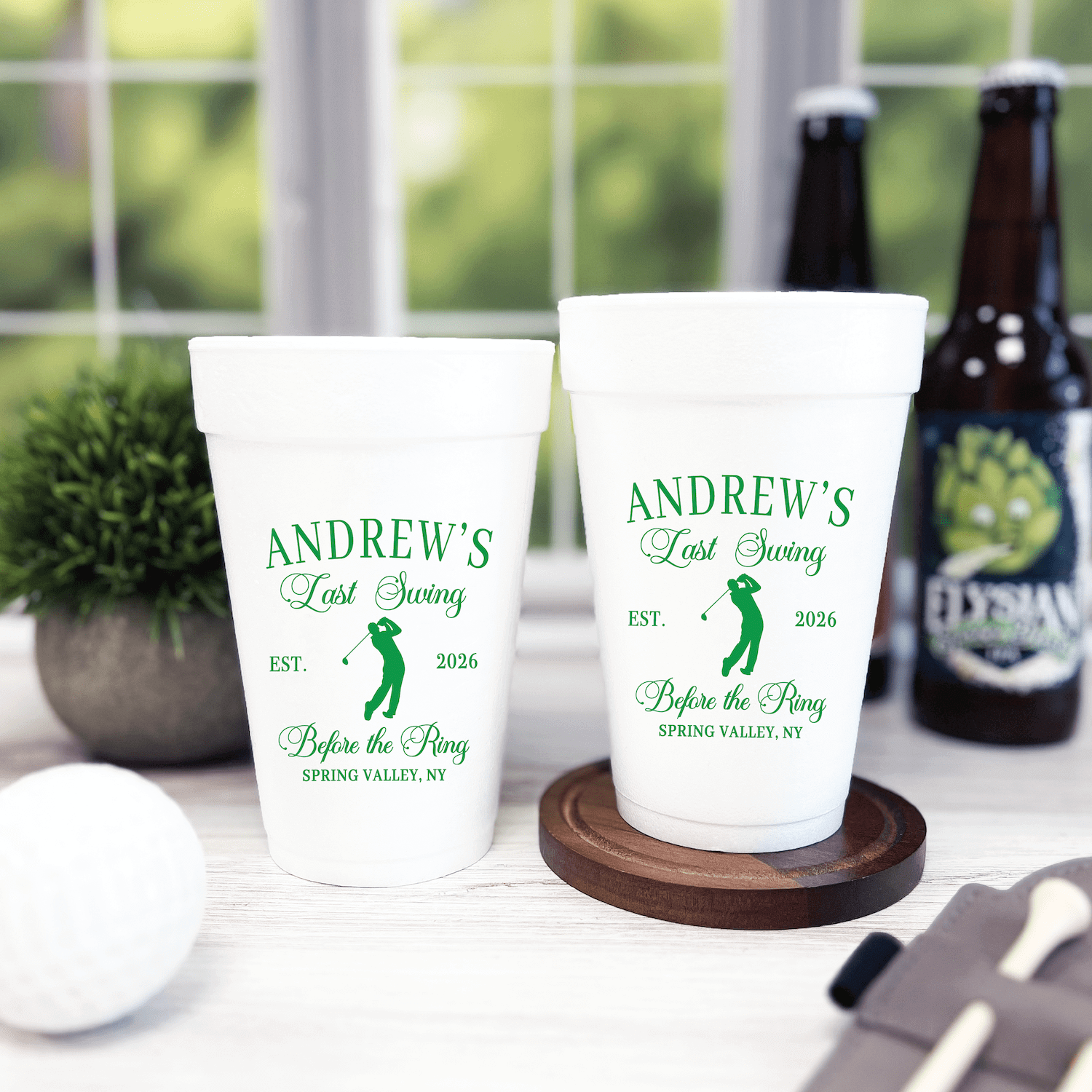 White foam cups personalized with "Andrew's Last Swing Before The Ring" text and golf design in green, displayed with beer bottles and wooden coaster