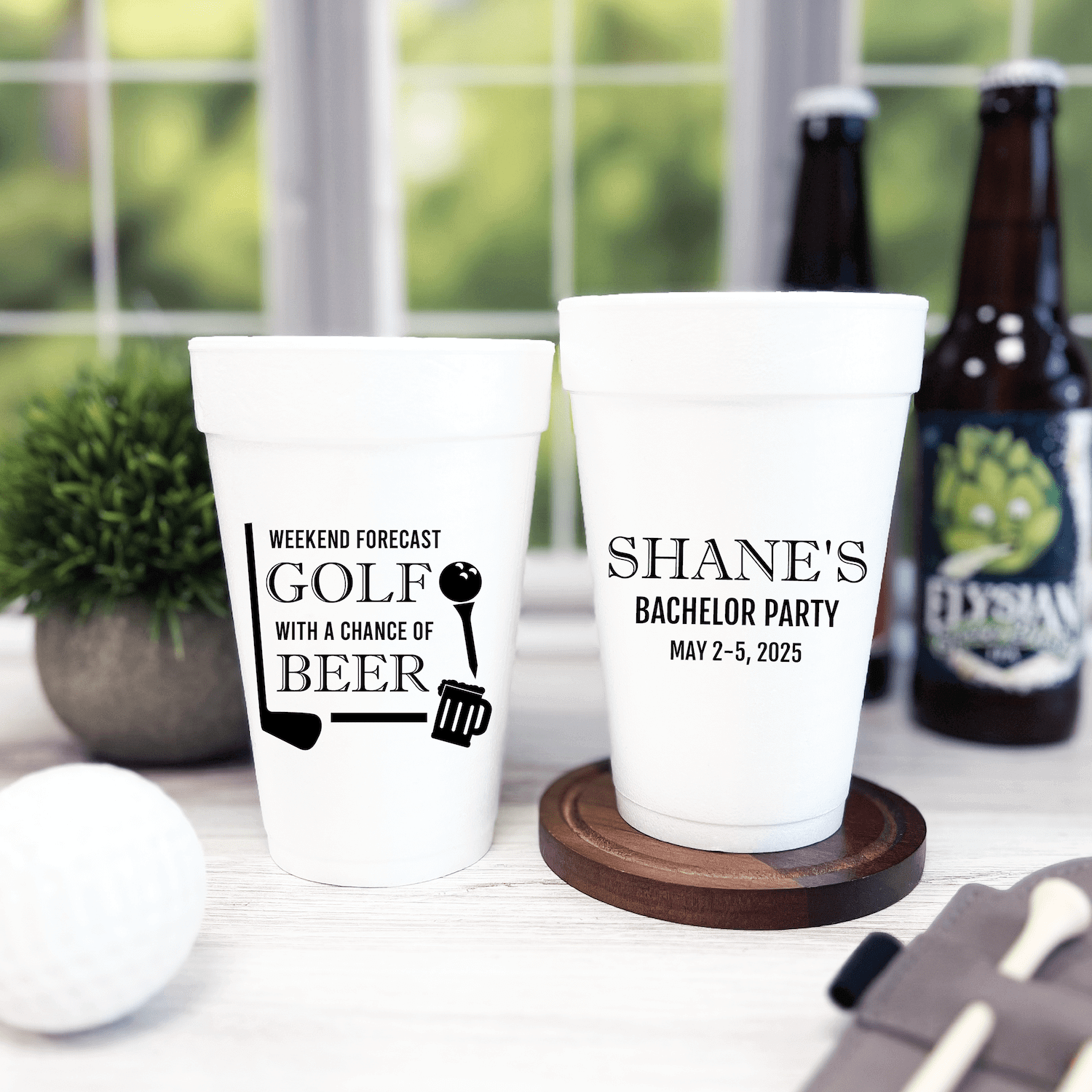 Two white foam cups with black personalized text, one reading "Weekend Forecast Golf with a Chance of Beer" and the other "Shane's Bachelor Party May 2-3, 2025" displayed with beer bottles and greenery.