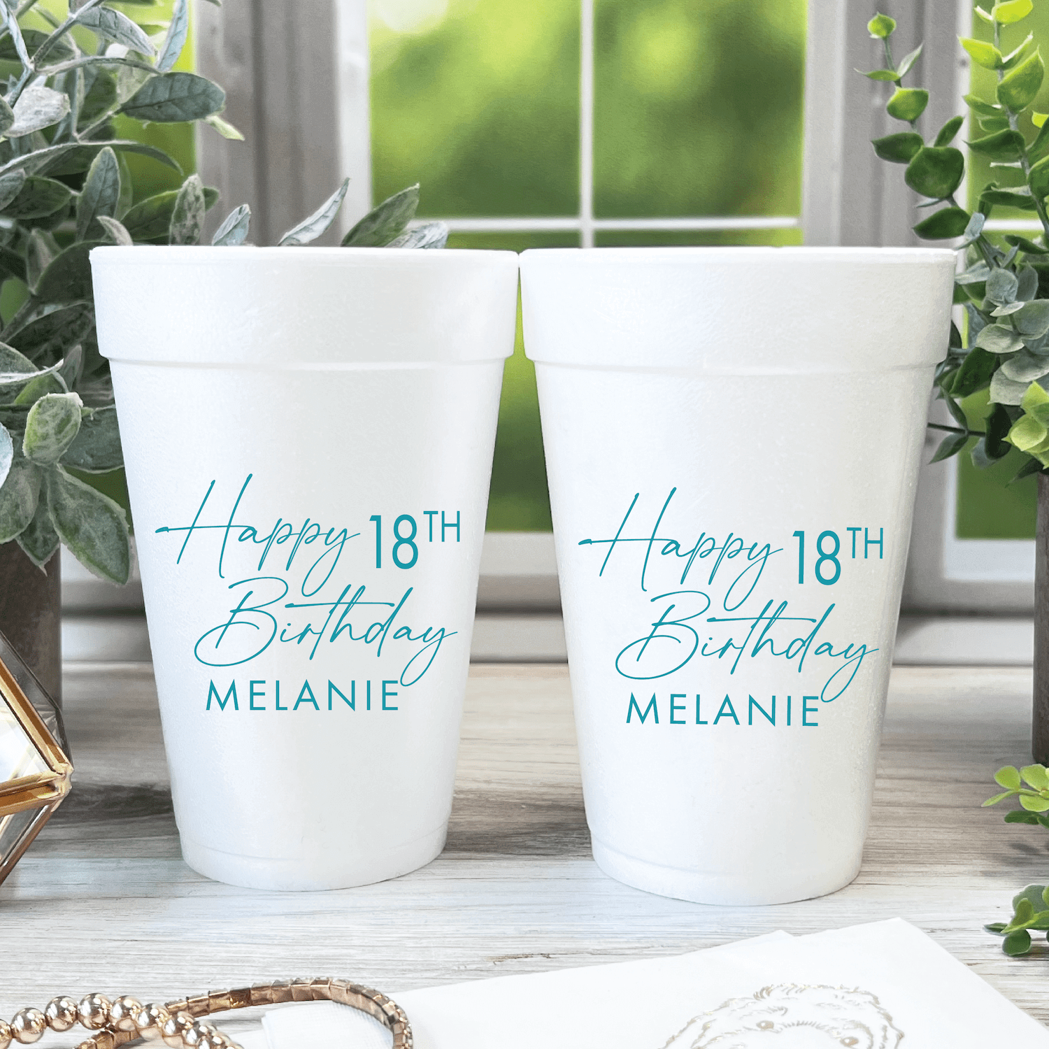 Two white foam cups personalized with "Happy 18th Birthday Melanie" in blue script text, displayed on a windowsill with greenery.