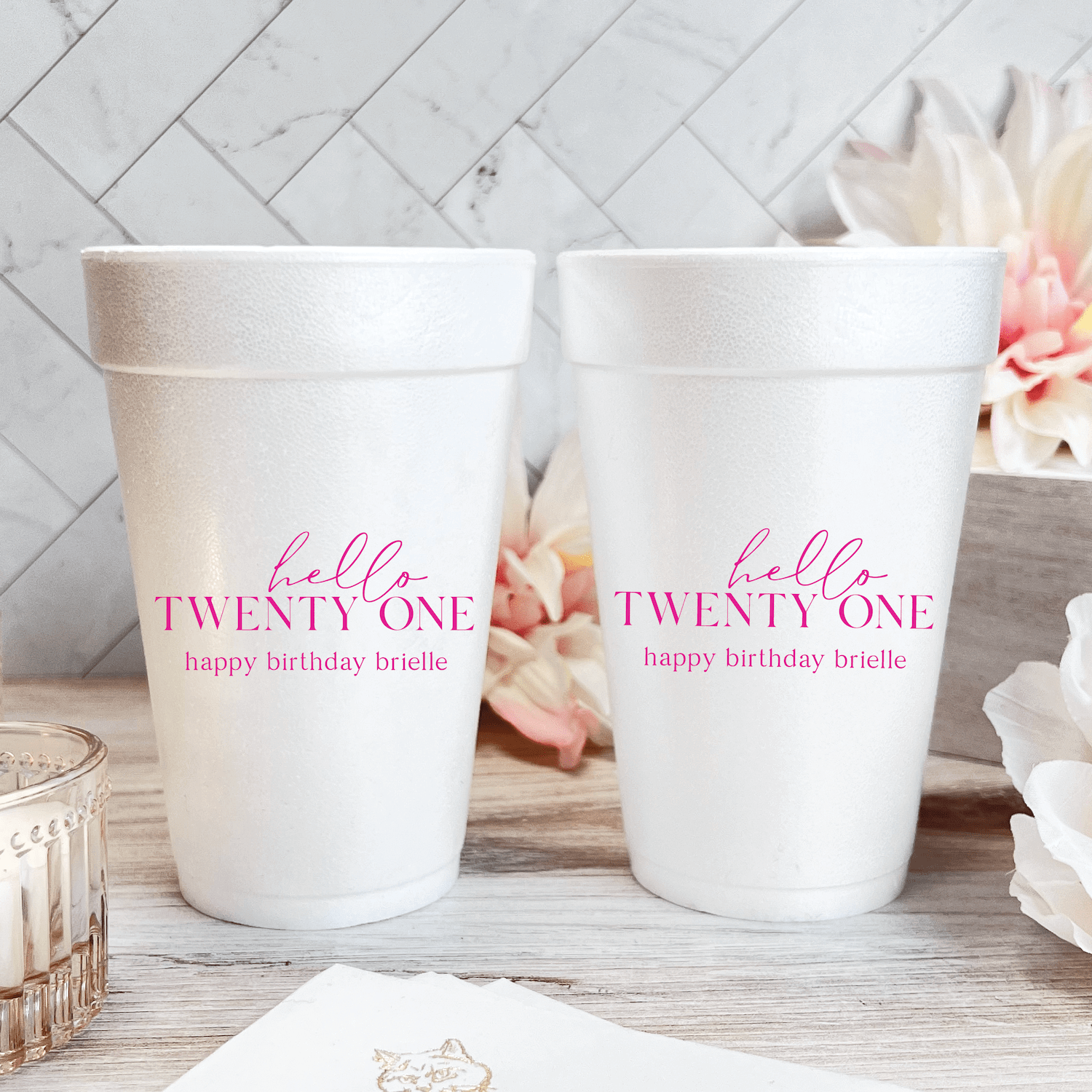 Two white foam cups with pink "hello TWENTY ONE" text and "happy birthday brielle" personalization displayed on white wood surface with flowers.