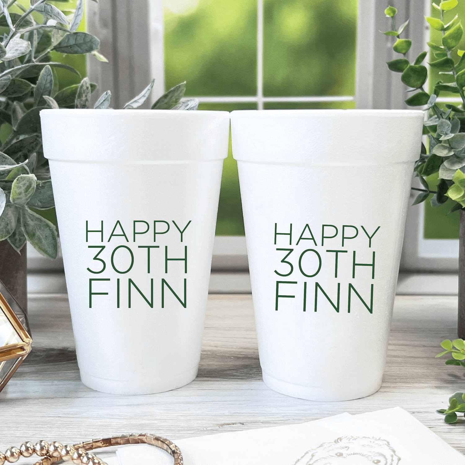 Two white foam cups printed with "HAPPY 30TH FINN" in gray text, displayed on a windowsill with greenery