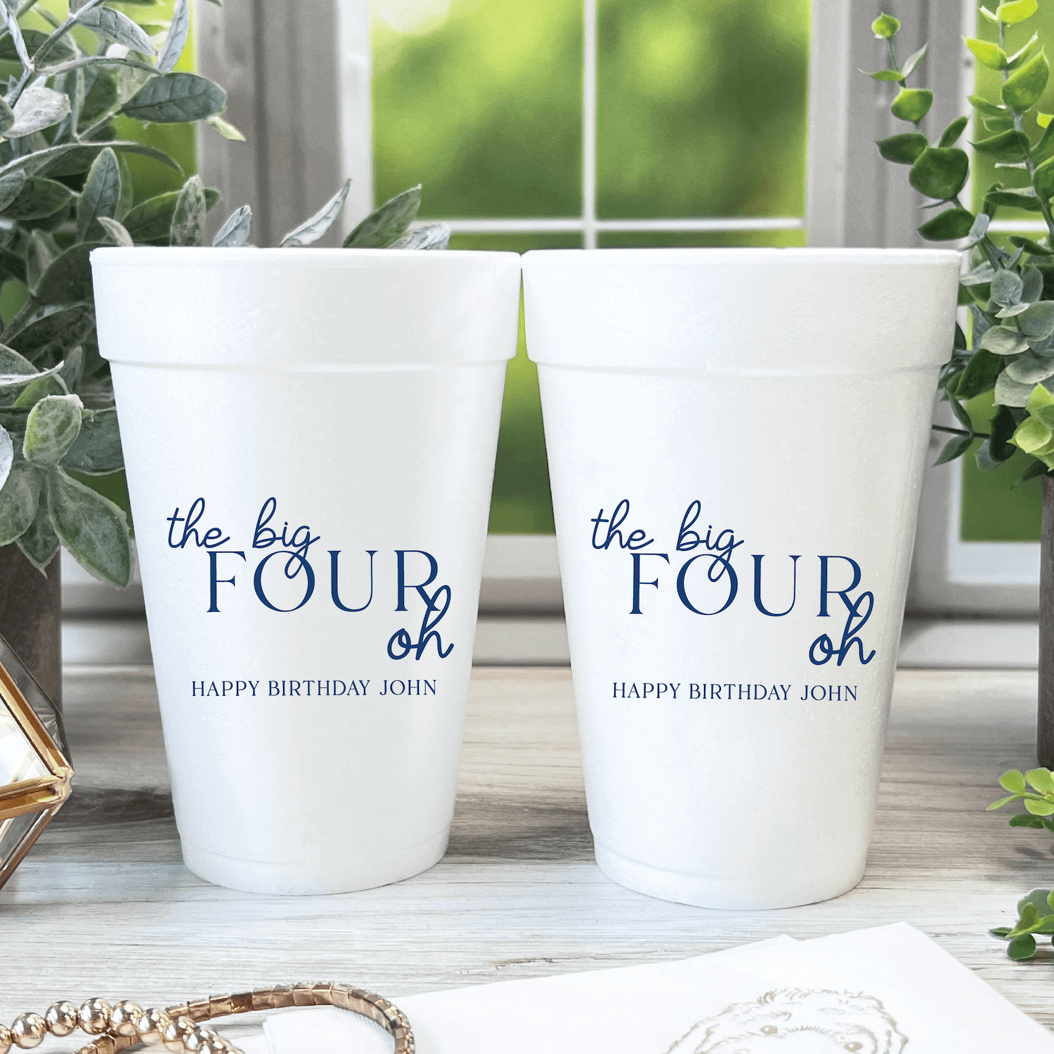 Two white foam cups printed with "the big FOUR oh" and "HAPPY BIRTHDAY JOHN" in navy blue text, displayed on a windowsill with greenery.
