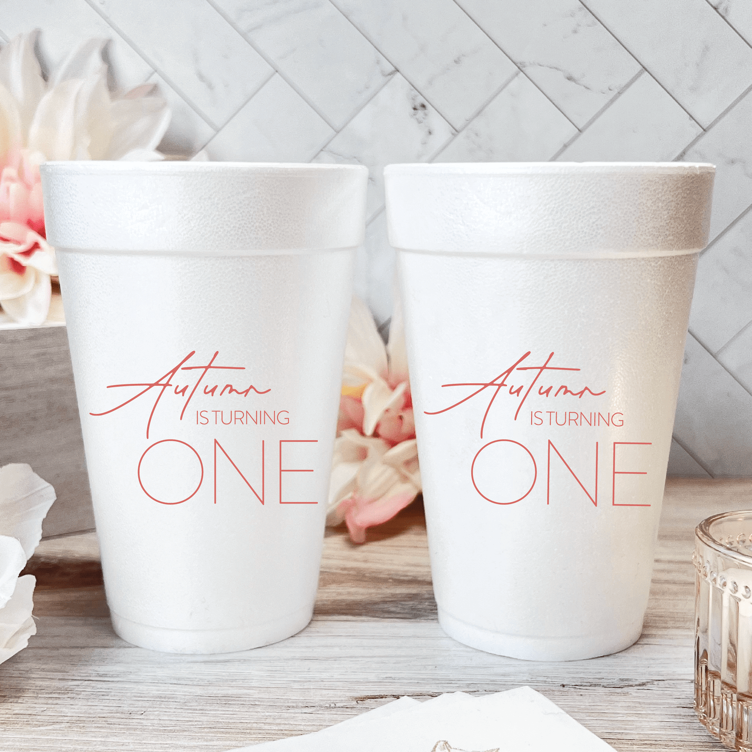 Two white foam cups with "Autumn is turning ONE" text in rose gold lettering on a marble tile background with floral accents