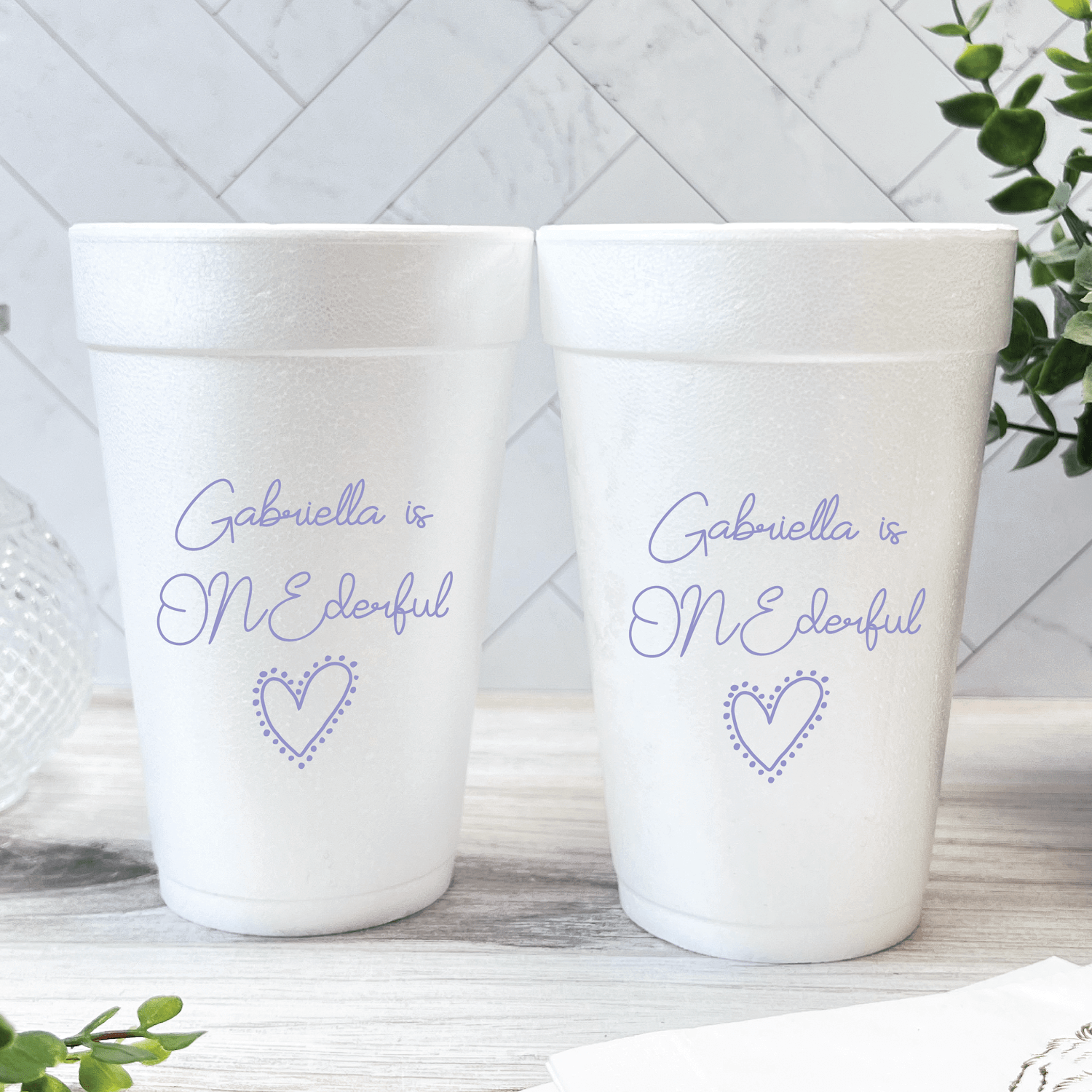 Two white foam cups personalized with "Gabriella is ONEderful" text and heart design in purple script