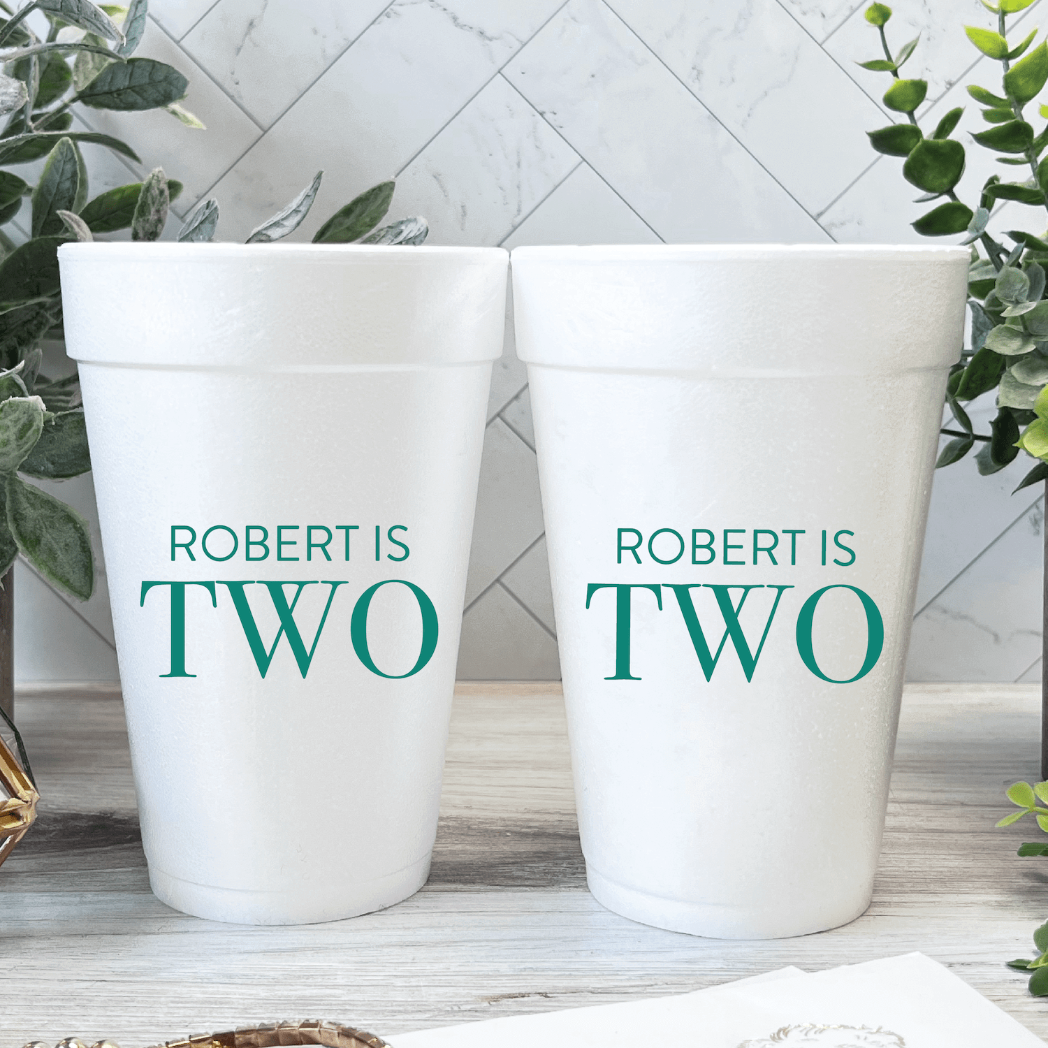 Two white foam cups with teal text reading "ROBERT IS TWO" displayed on herringbone tile background with green plants.