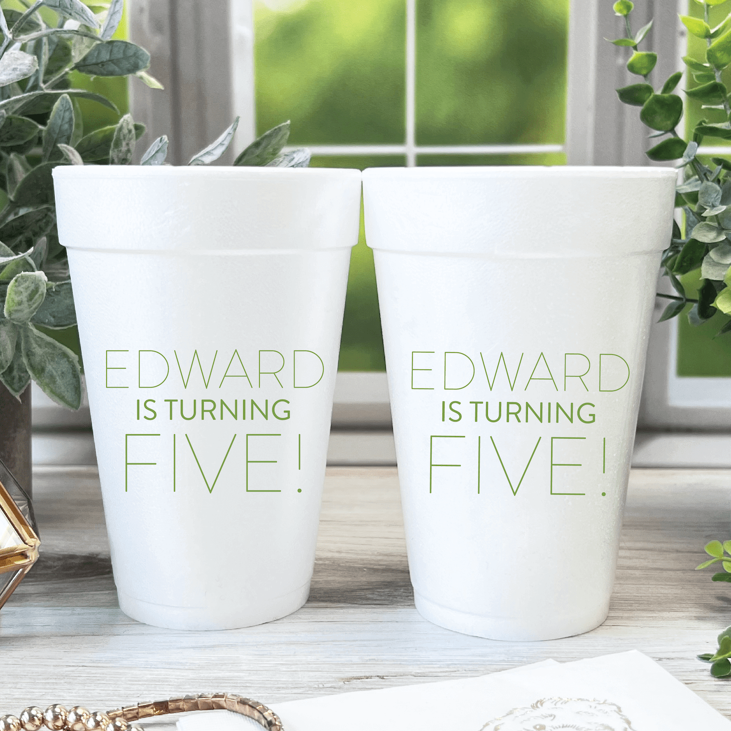 Two white foam cups with sage green text reading "EDWARD IS TURNING FIVE!" displayed on a wooden surface near a sunny window with plants.