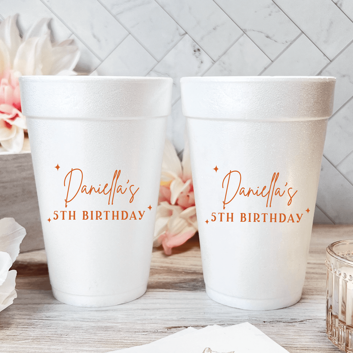 White foam cups with rose gold "Danielle's 5th Birthday" personalization text, displayed in pairs on white wood surface with pink flowers