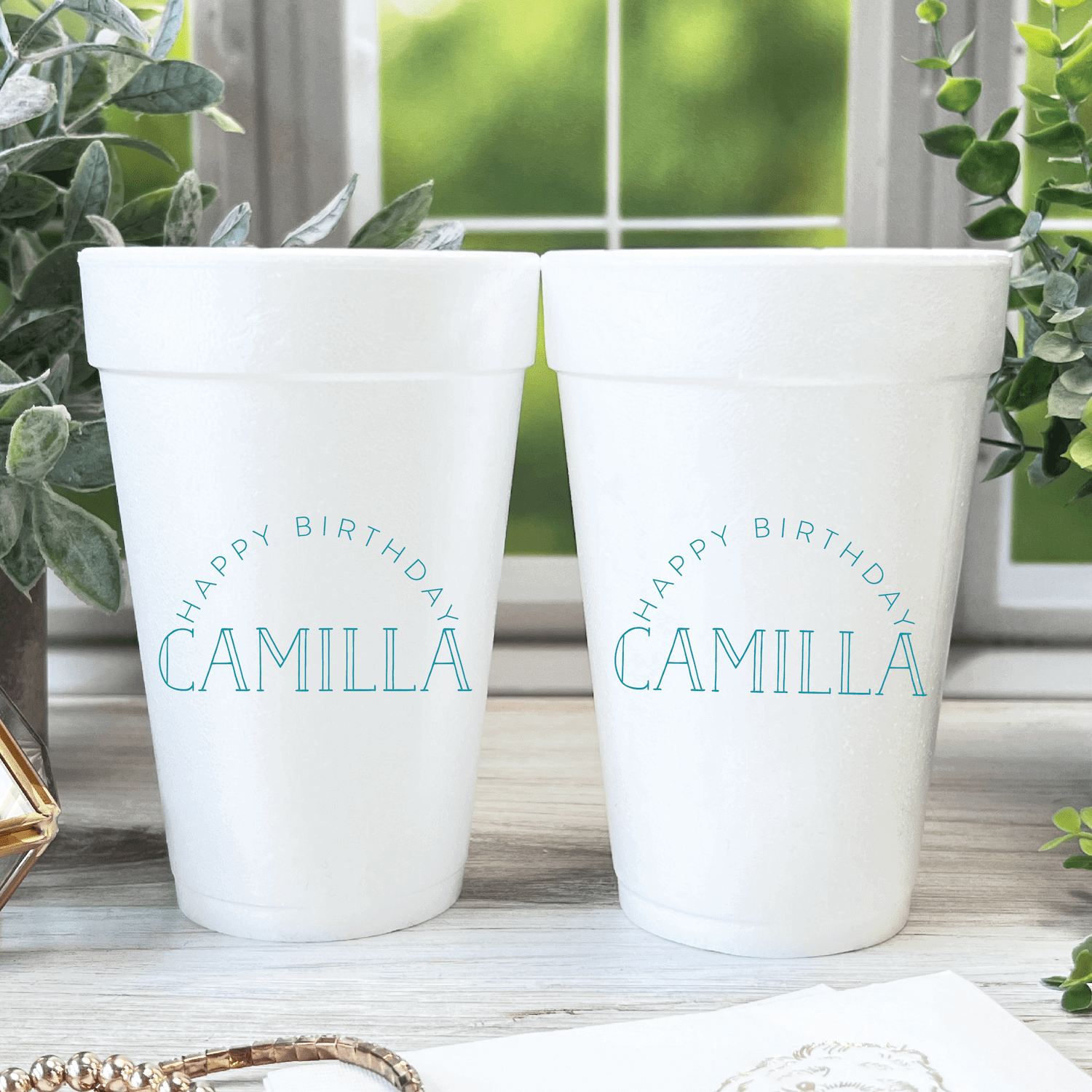 Two white foam cups personalized with "Happy Birthday Camilla" in blue text, displayed on a wooden surface near a window with plants.