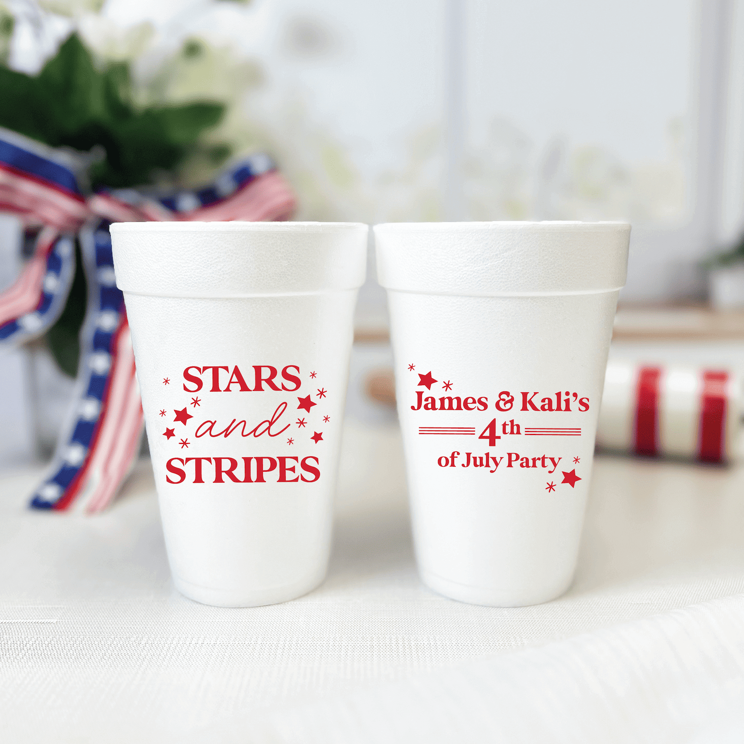 Two white foam cups with red personalized text reading "STARS and STRIPES" and "James & Kali's 4th of July Party" with star designs, displayed on a white surface with patriotic decorations.