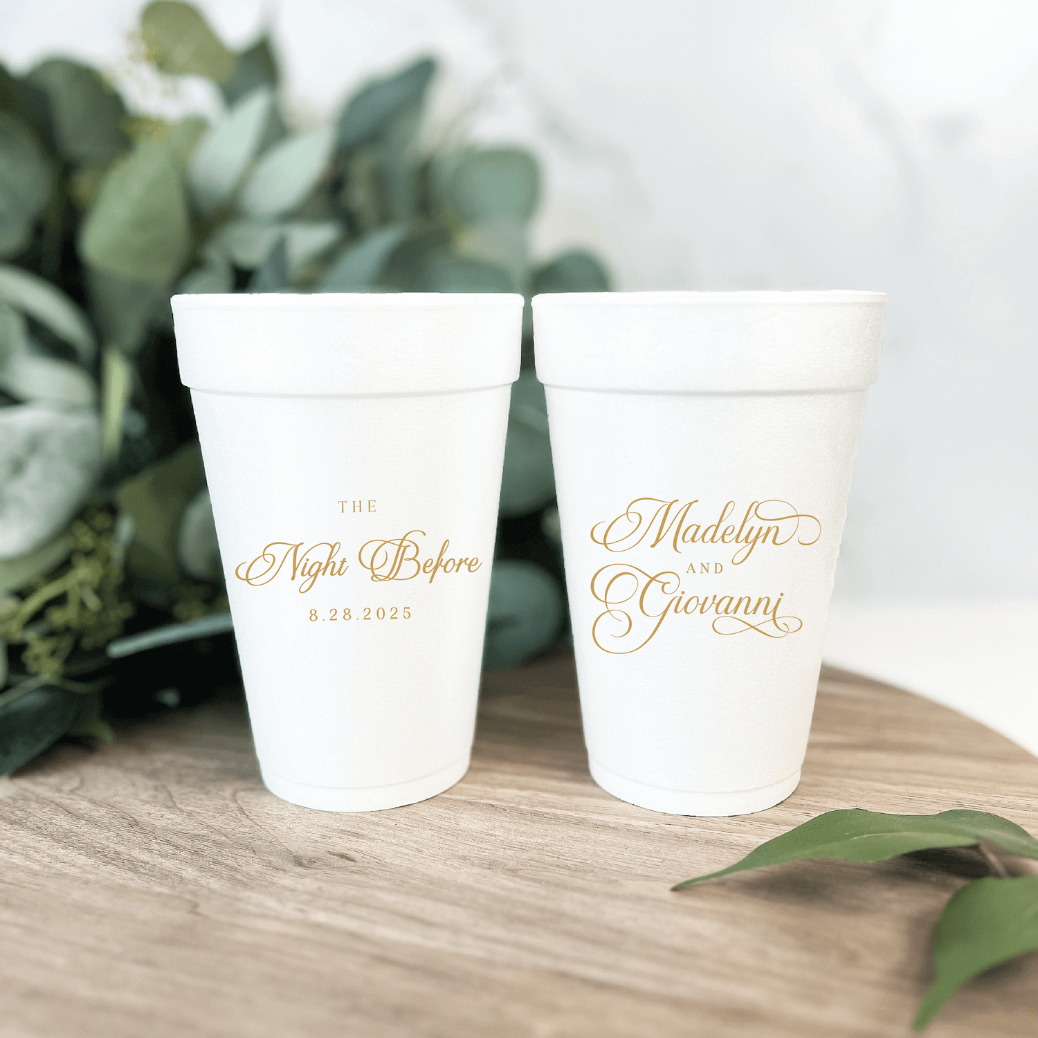 Two white foam cups with gold script text and dates, displayed on wood with greenery in background