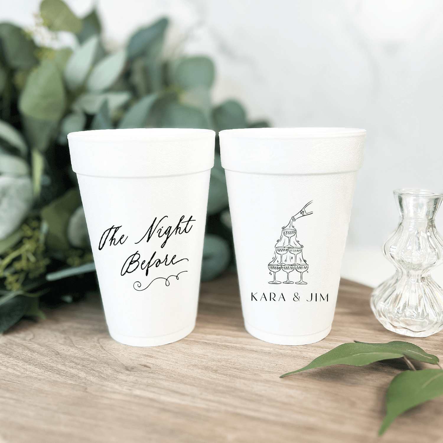 White foam cups with "The Night Before" script text and "KARA & JIM" with cake design, displayed with greenery and glassware.