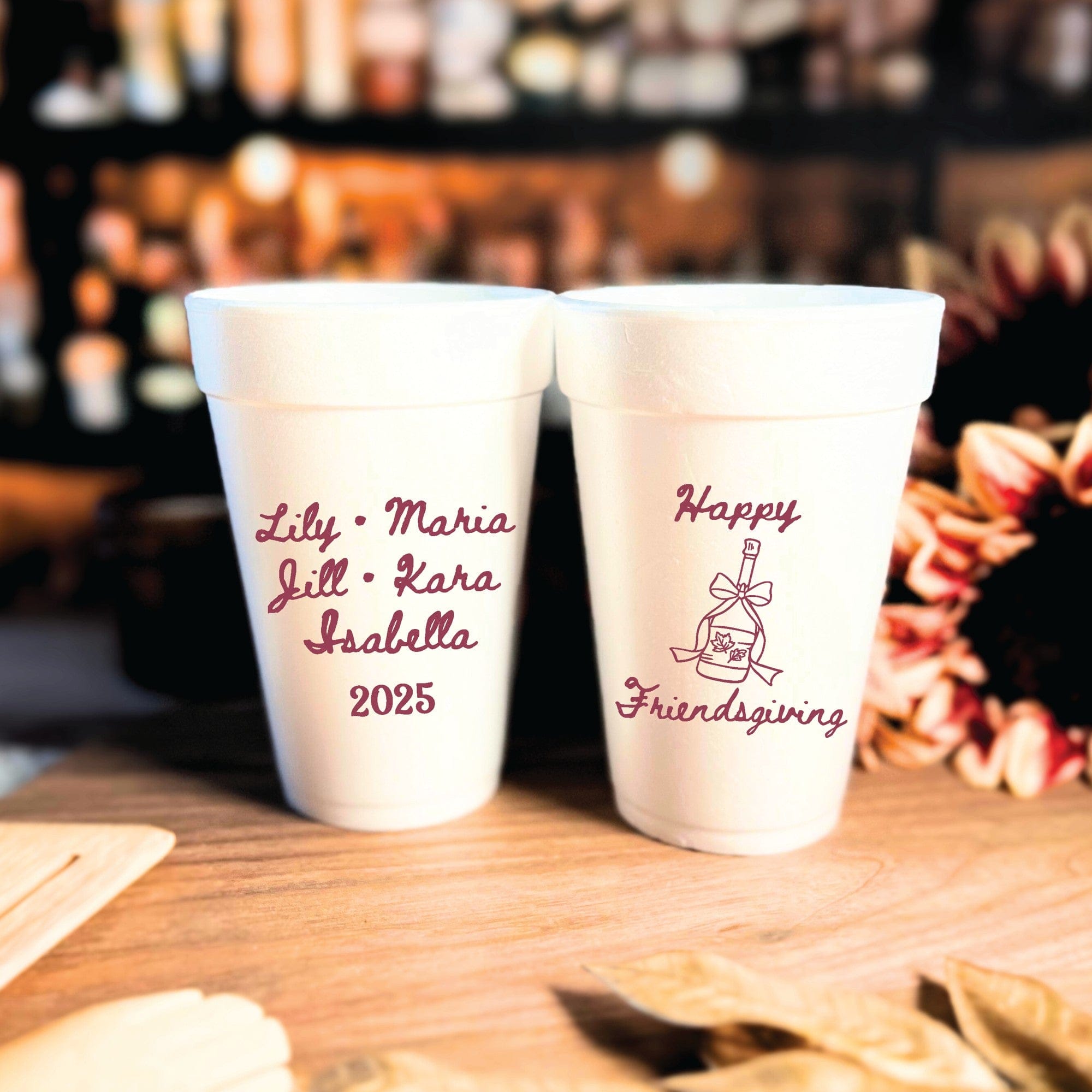 Two white foam cups feature dark red personalized designs, one with custom text and the other with a festive fall bottle illustration, ideal for a Friendsgiving or autumn gathering.