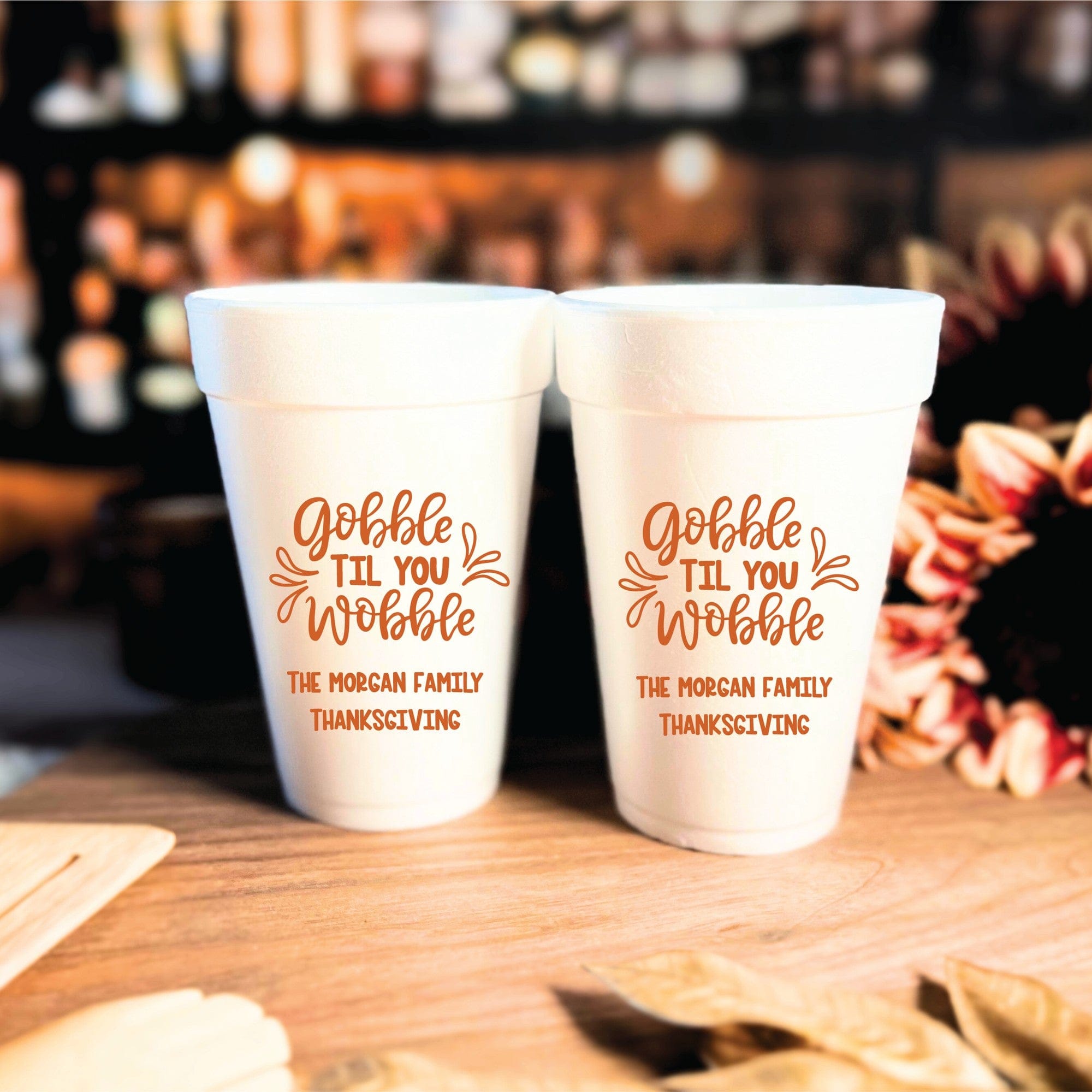 Two white foam cups with orange custom Thanksgiving-themed printing are displayed on a wooden surface for an event.