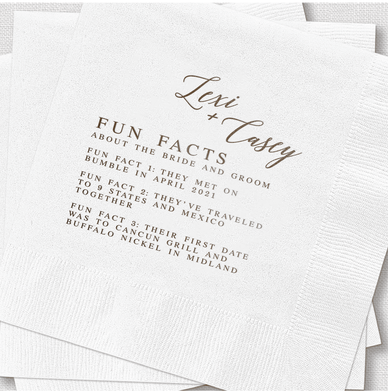 White napkin with gold foil stamped text reading "Lexi + Tash" and "Fun Facts About the Bride and Groom" with personalized wedding details listed below.
