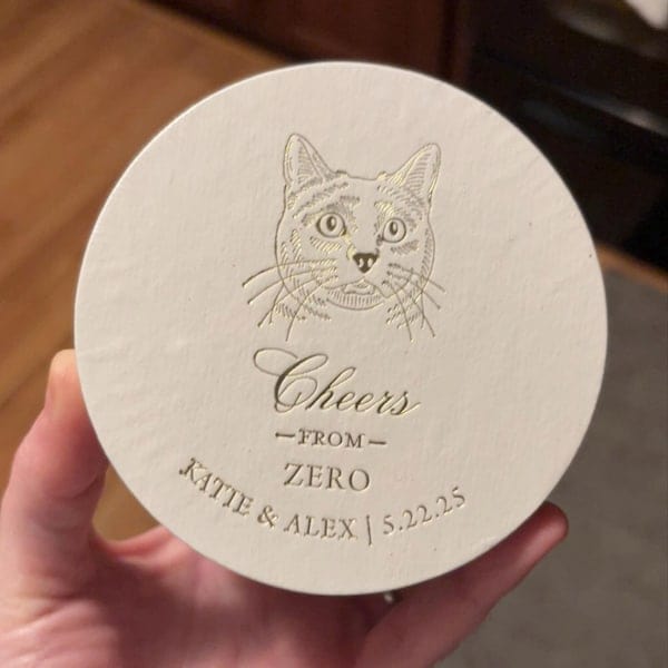 Cream-colored round coaster with hand-drawn cat illustration, "Cheers from Zero" text, and personalized names "Katie & Alex" with date 5.22.25