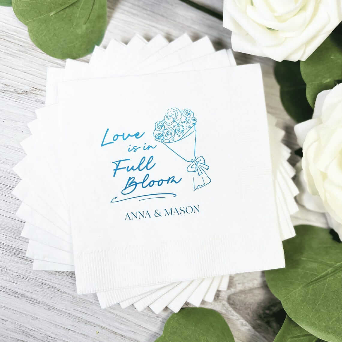 White napkin with blue foil-stamped design showing watering can and "Love is in Full Bloom Anna & Mason" text