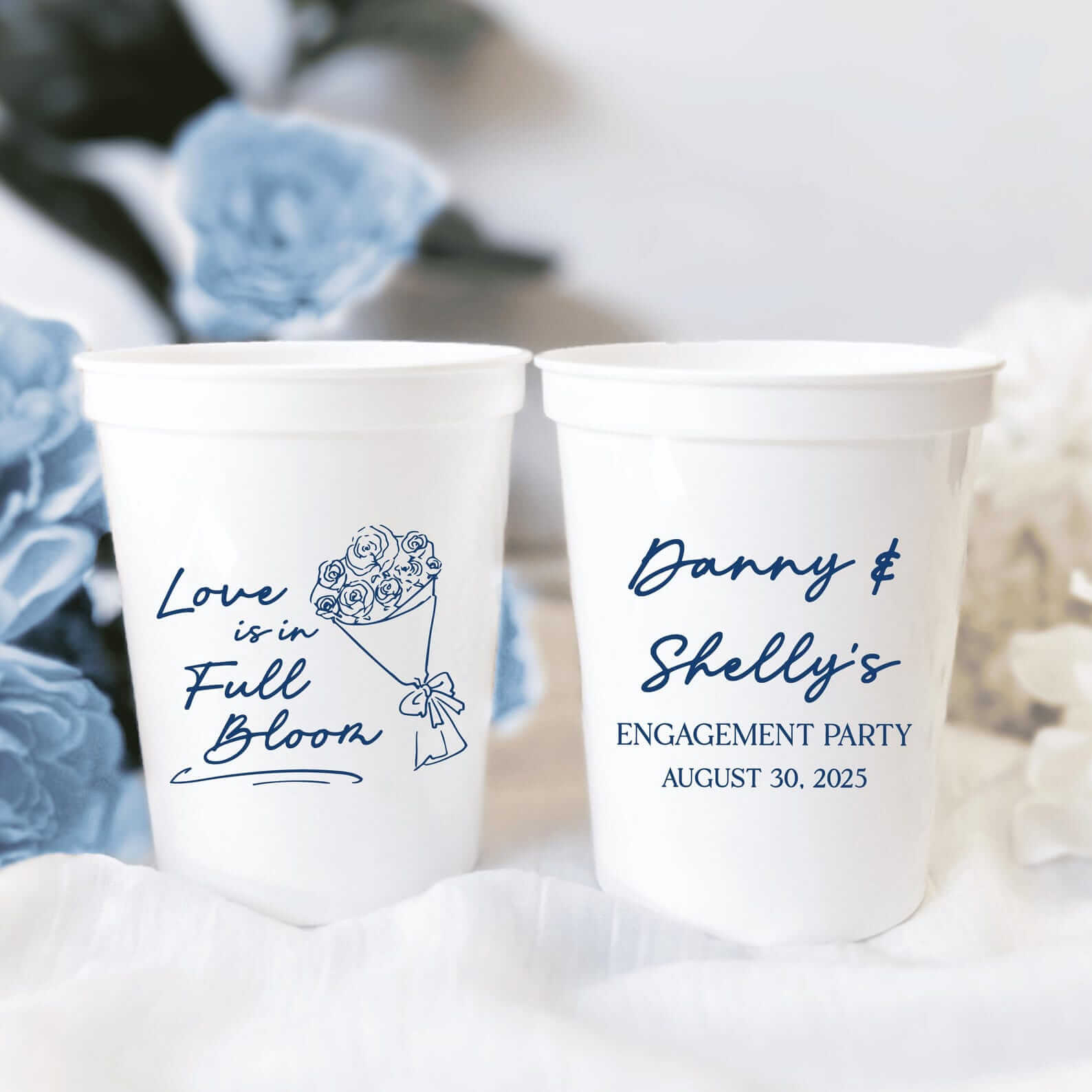 Two white stadium cups with navy blue text and floral designs, one reading "Love is in Full Bloom" and the other personalized "Danny & Shelly's Engagement Party August 30, 2023"