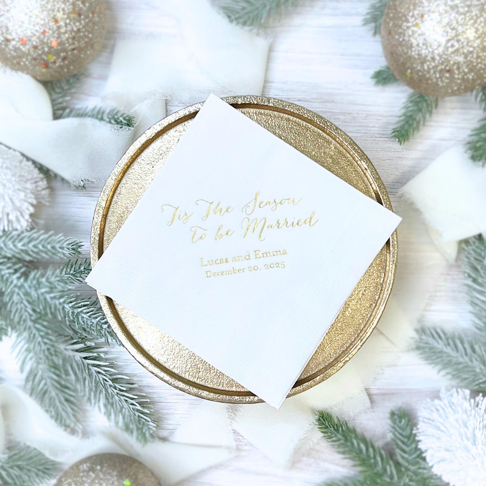 White cocktail napkin with gold foil text "Tis the Season to be Married" on gold charger plate, surrounded by holiday greenery and ornaments