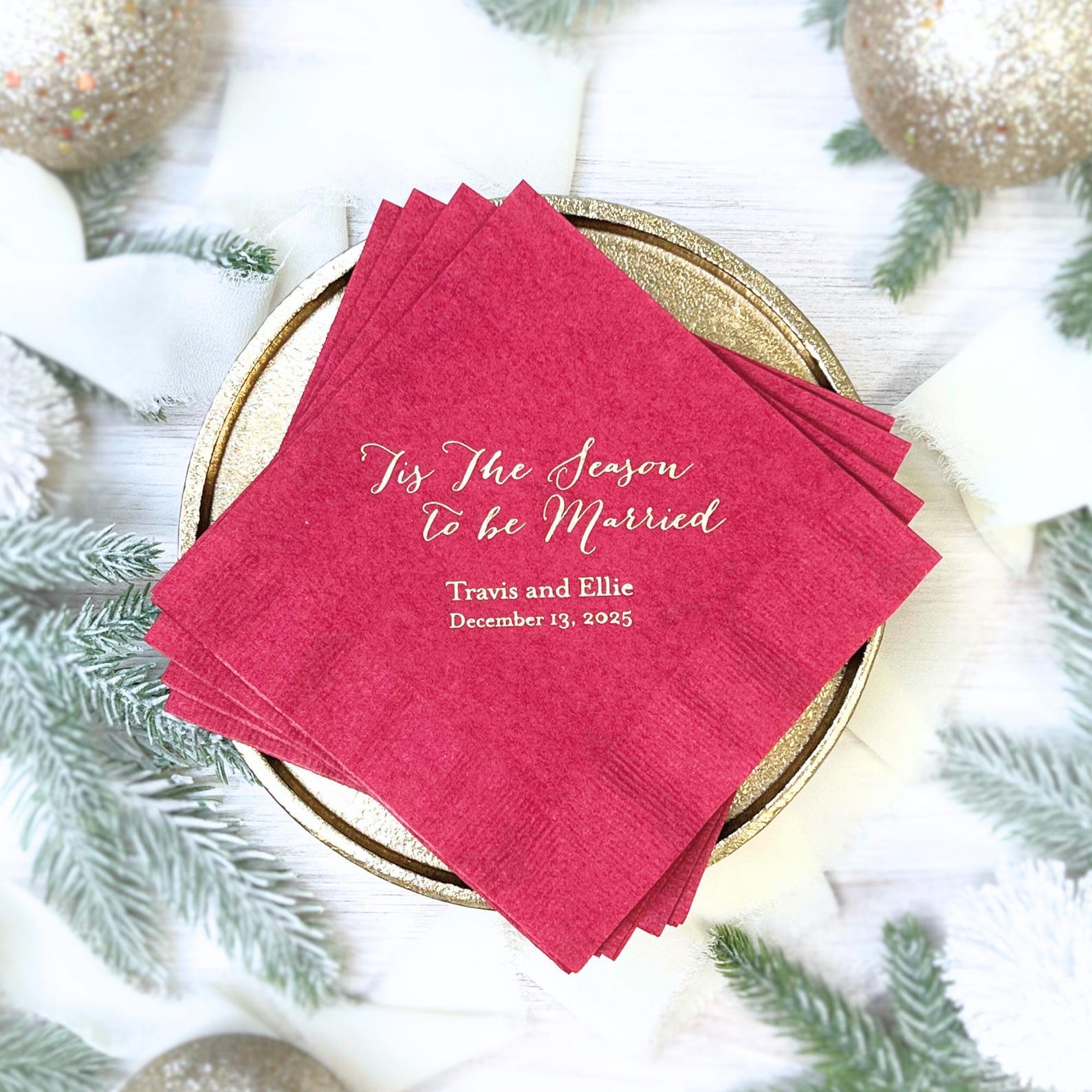 Hot pink napkins with gold foil text reading "Tis the Season to be Married" and couple names and date, arranged on gold charger plate.