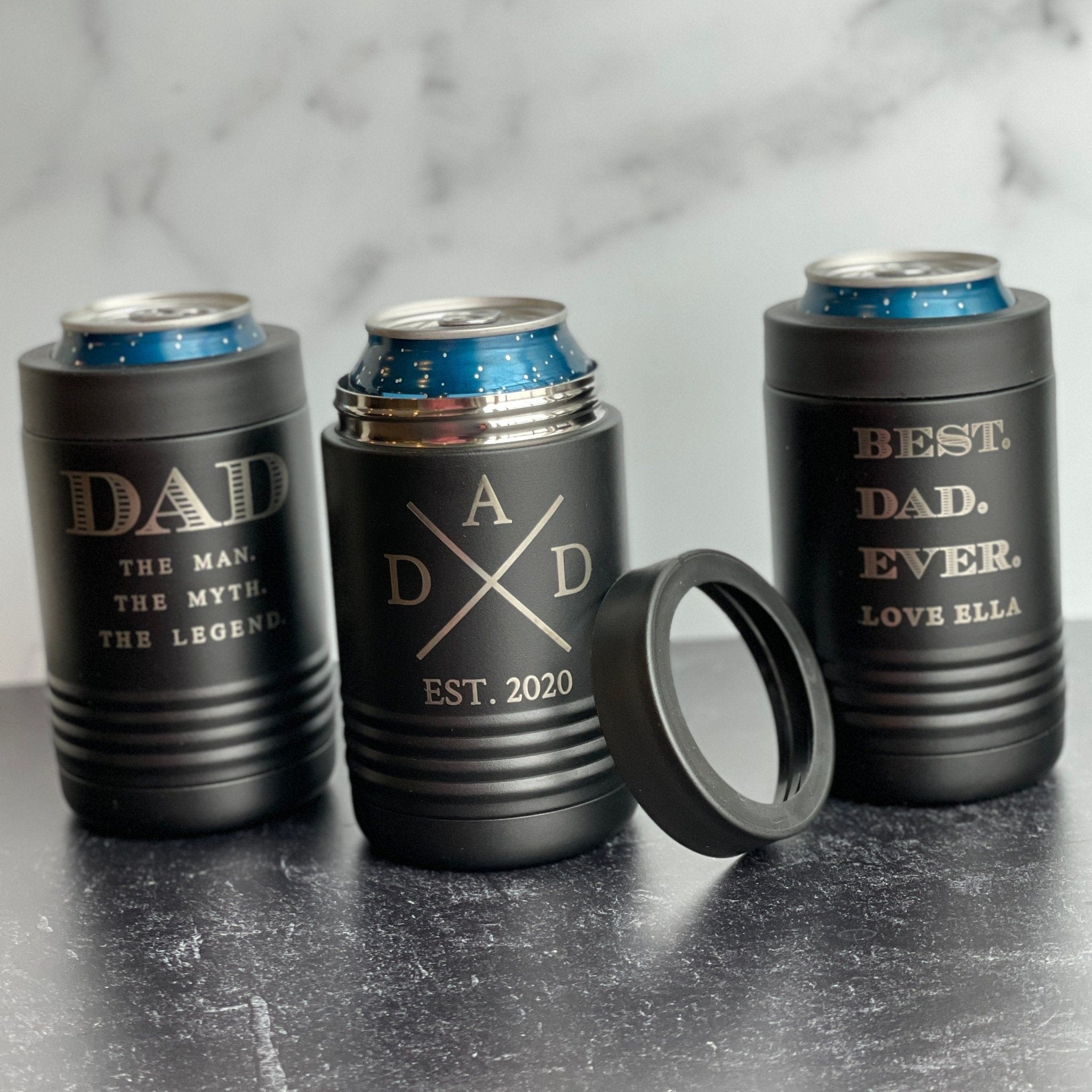 Three black metal can coolers with gold engraved text reading "Dad the Man the Myth the Legend," "Dad Est. 2020," and "Best Dad Ever Love Ella," displayed with blue cans and a detachable ring on marble surface.