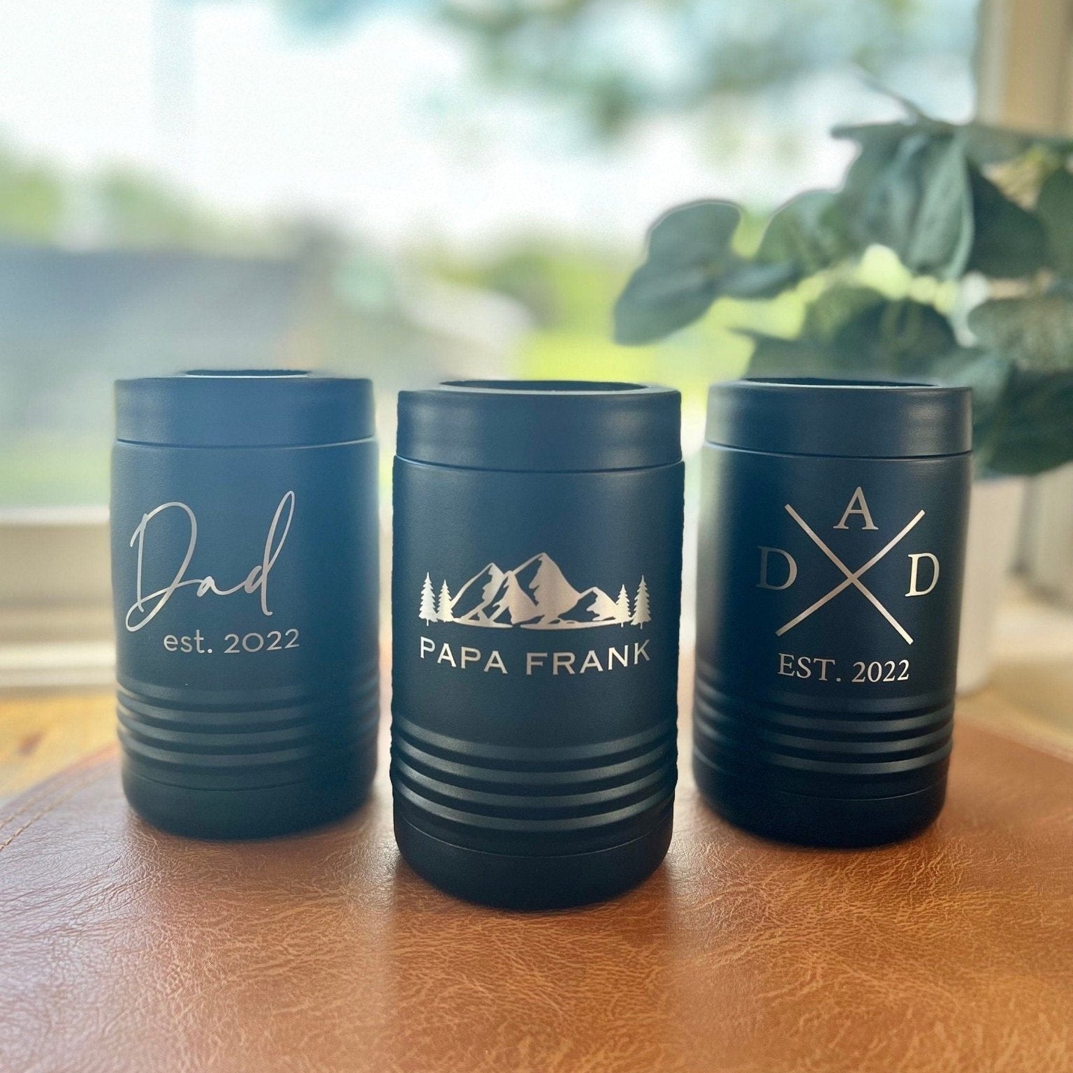 Three navy metal can coolers with personalized text including "Dad est. 2022," "Papa Frank," and monogram design with establishment date, displayed on wooden table by window