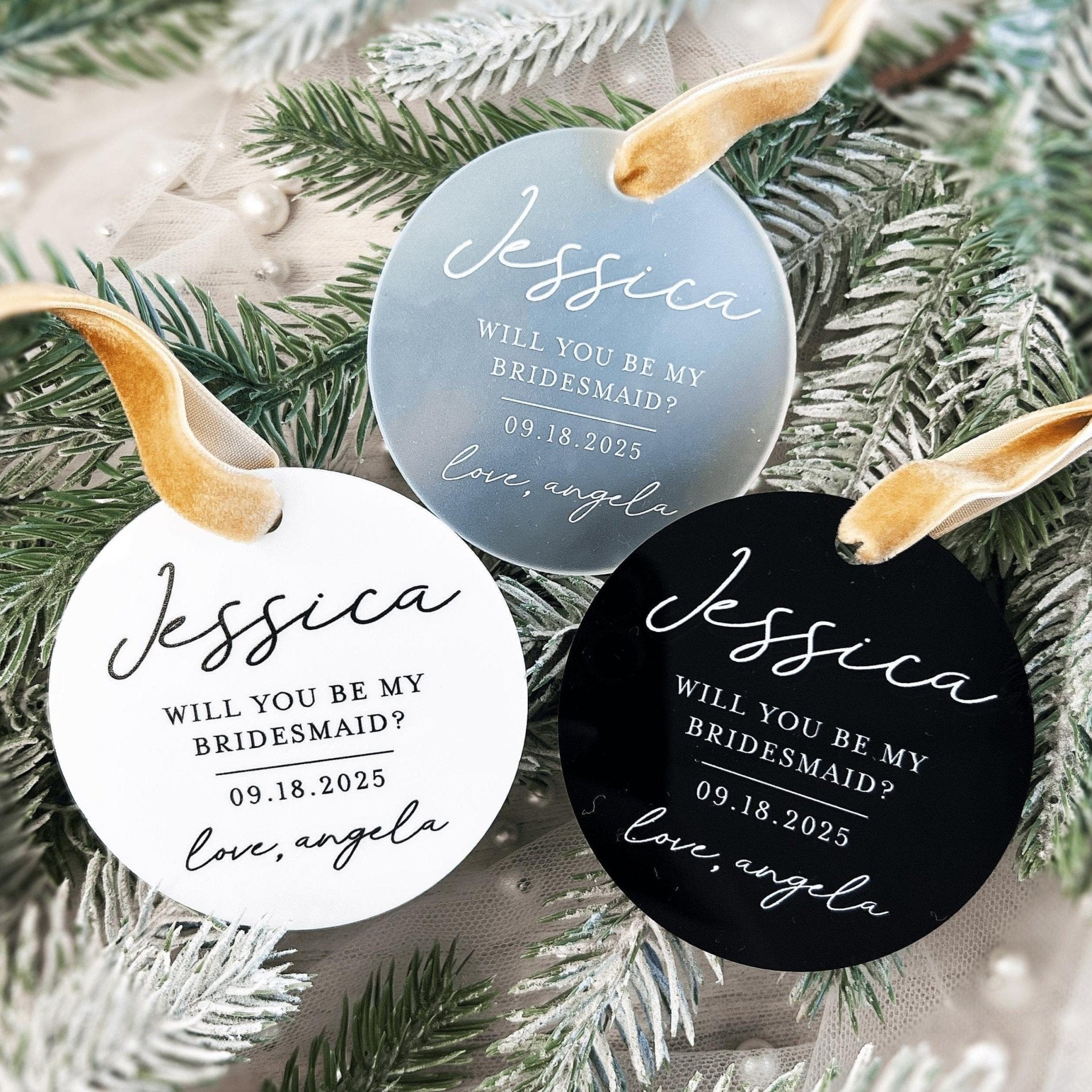 Three personalized acrylic ornaments in blue, white, and black with gold ribbon, each printed with "Will you be my bridesmaid?" and a name and date.
