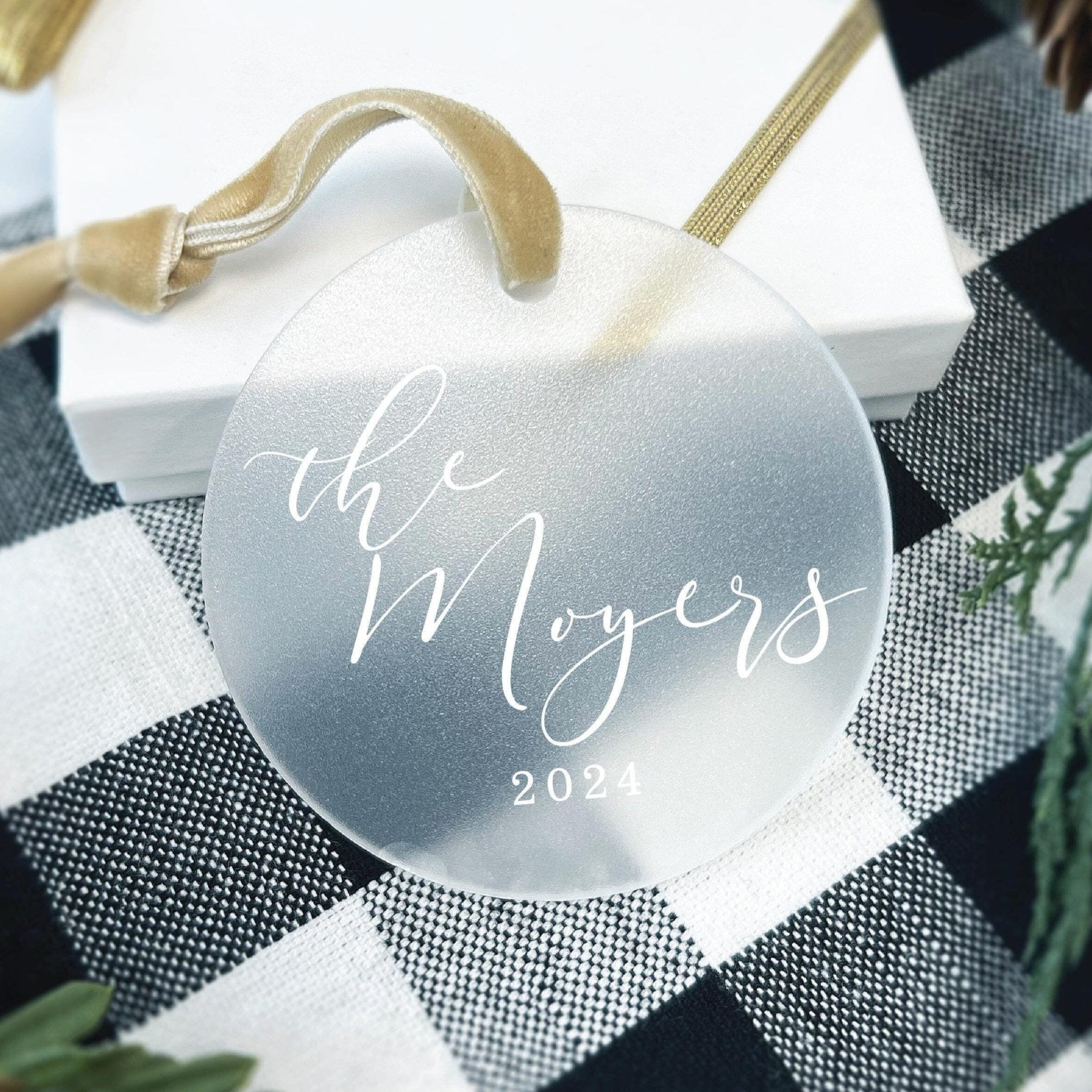 Frosted acrylic round ornament with "the Moyers 2024" in script text and gold ribbon loop handle.