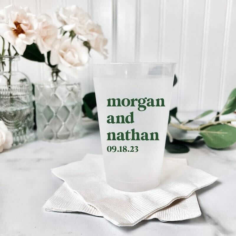 Frosted shatterproof cup printed with "morgan and nathan 09.18.23" in green text, displayed on white napkin with flowers in background.