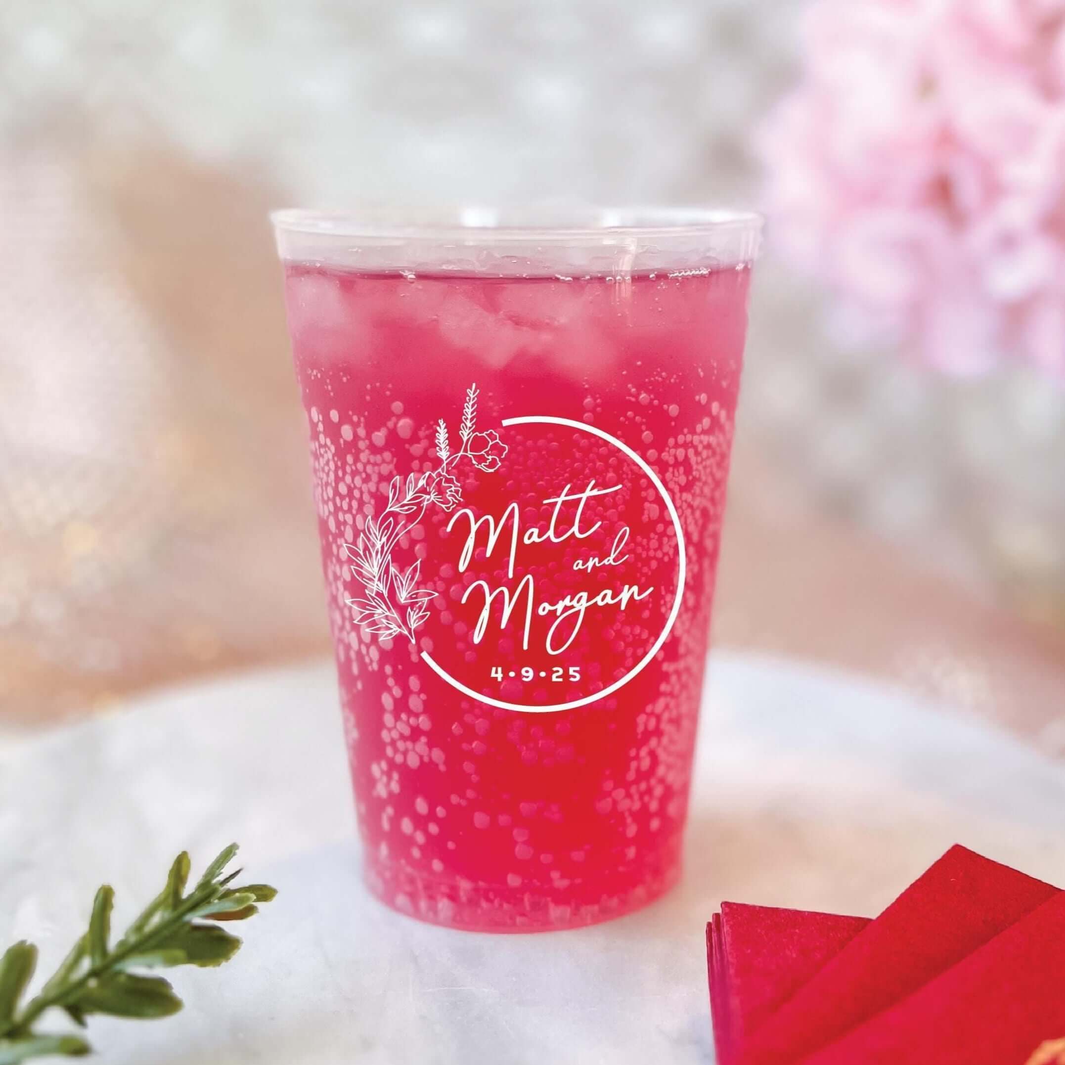 Red frosted cup filled with pink beverage, personalized with "Matt & Megan" and wedding date in white script lettering inside a circular floral wreath design.