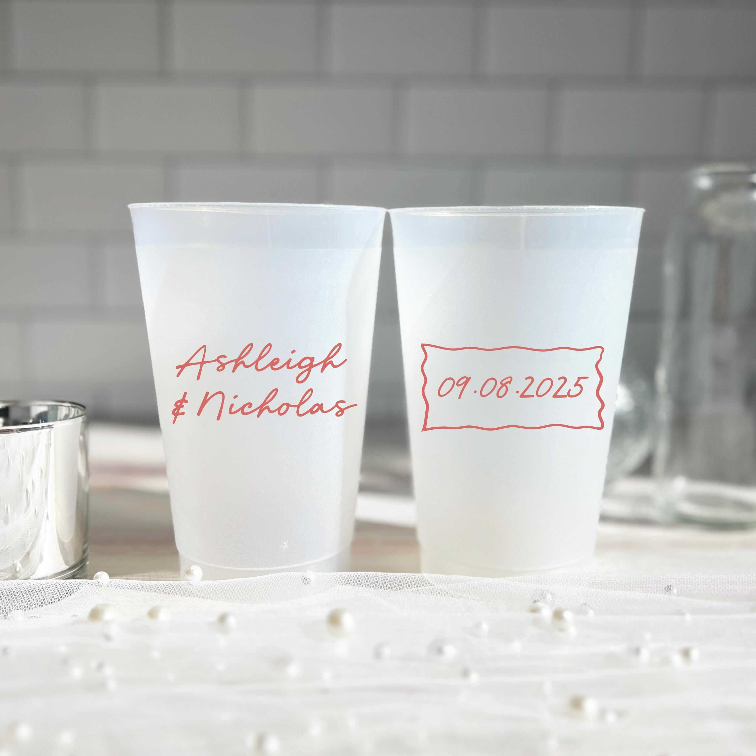 Two frosted shatterproof cups with red personalized text reading "Ashleigh & Nicholas" and "09.08.2025" displayed against a white subway tile background.