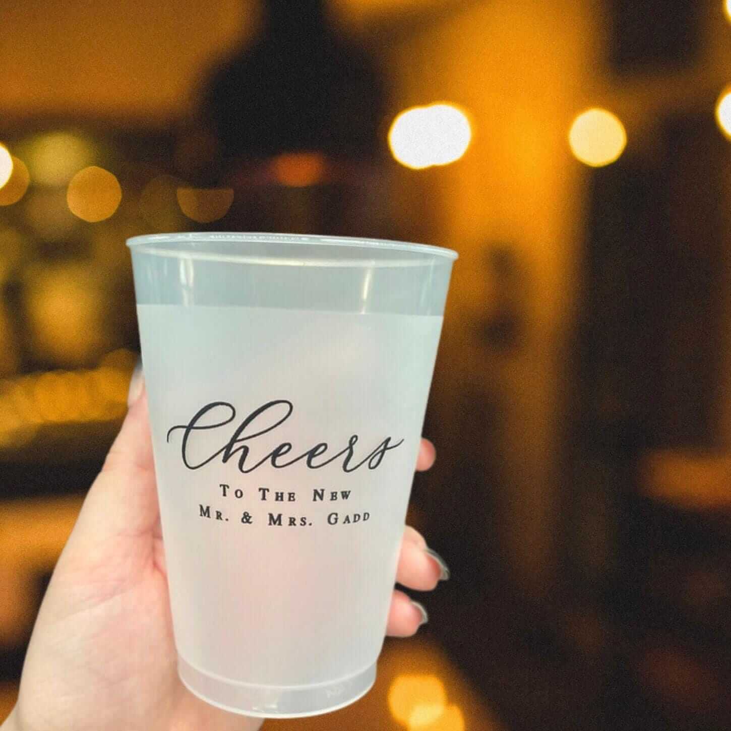 Frosted cup printed with "Cheers To The New Mr. & Mrs. Gapp" held in hand against warm bokeh background