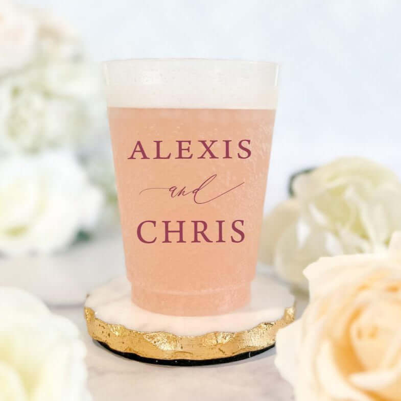 Frosted cup with "ALEXIS and CHRIS" personalized names in burgundy text, filled with rose-colored beverage, surrounded by white flowers.